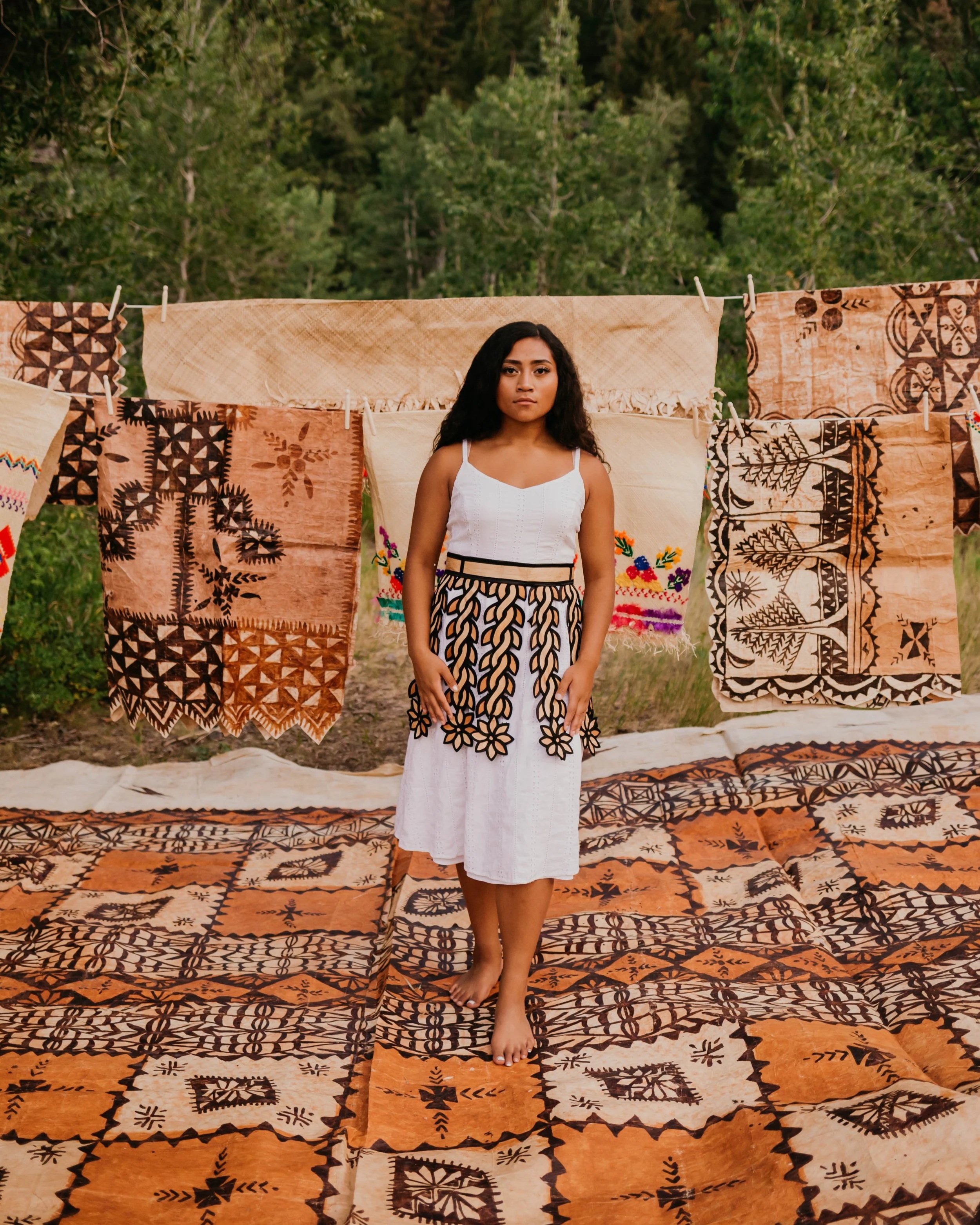 A woman standing barefoot on patterned textiles outdoors, with fabric and woven artwork hanging behind her, surrounded by green trees.