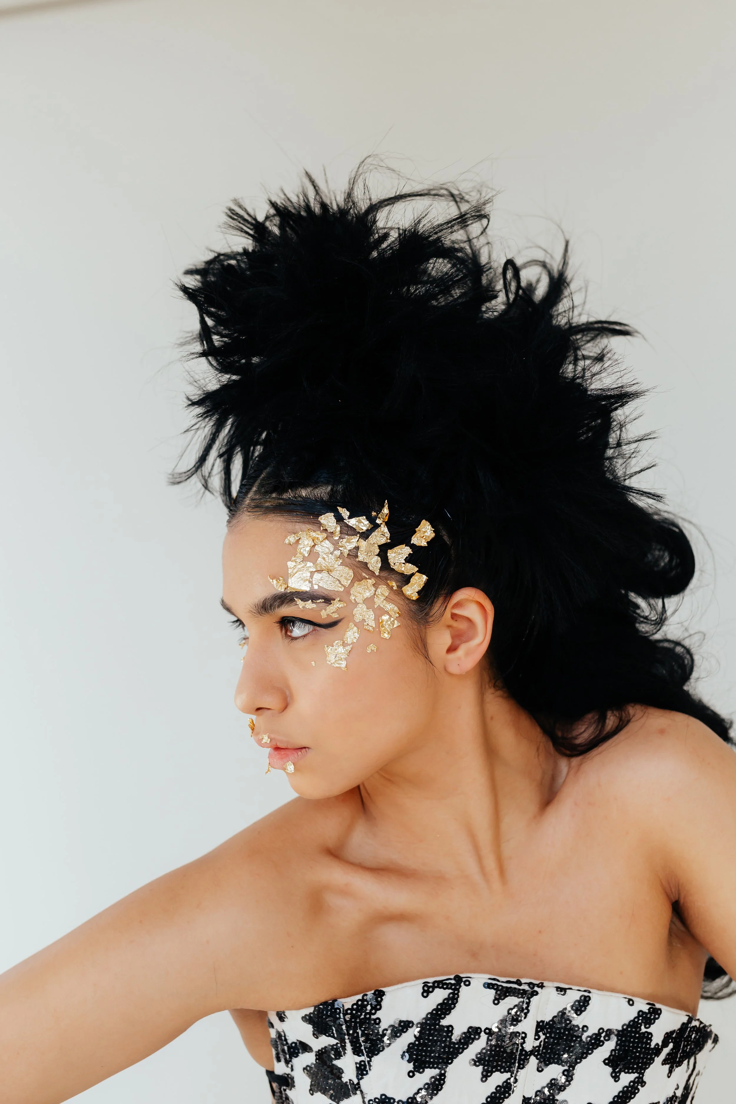 A woman with dramatic black hair styled into a voluminous, messy updo, wearing gold foil pieces on her face, and a strapless dress with a black and white pattern, against a plain white background.