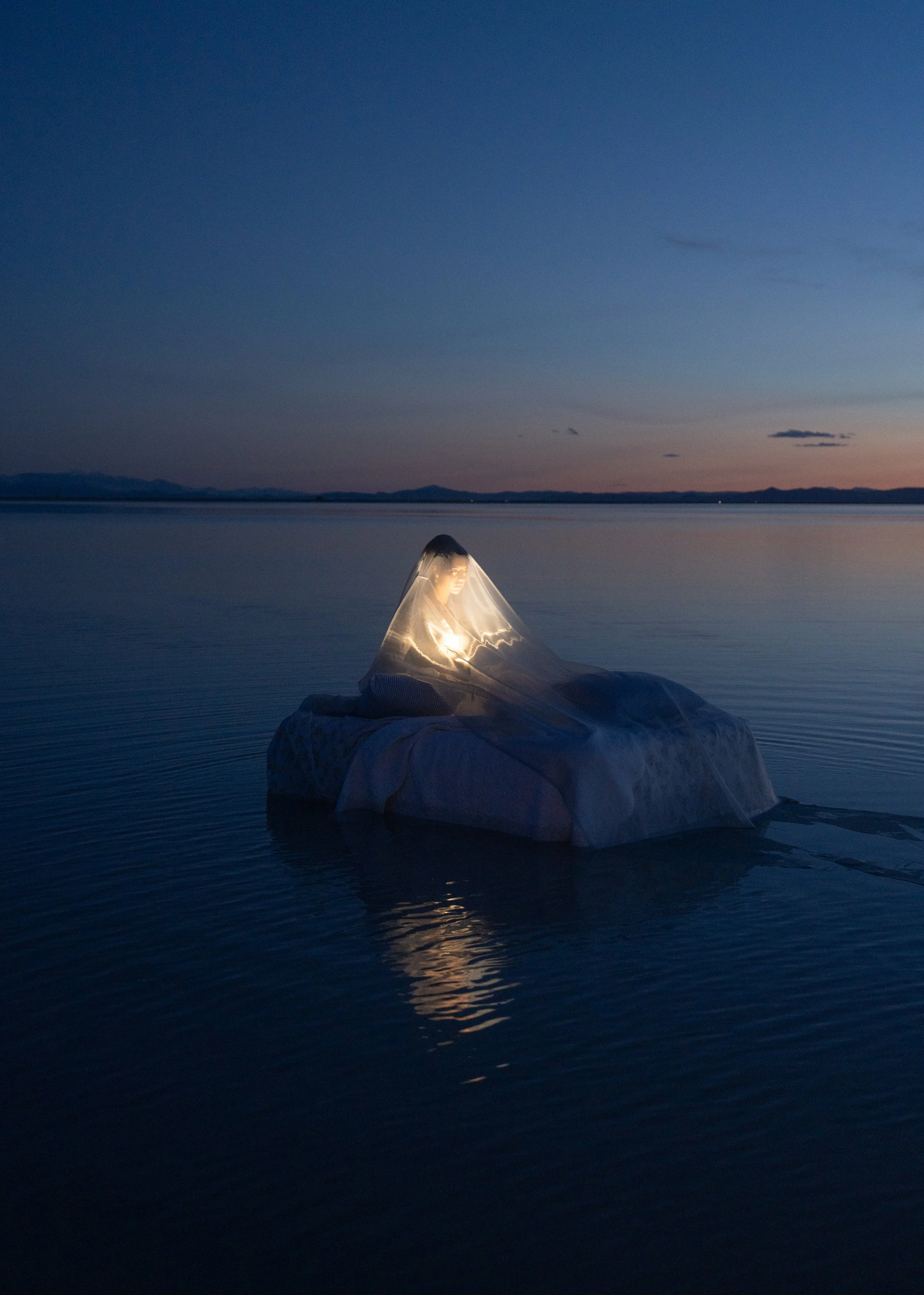 A person sitting on a boat in a calm body of water at dusk, covered with a transparent veil and holding a glowing light.