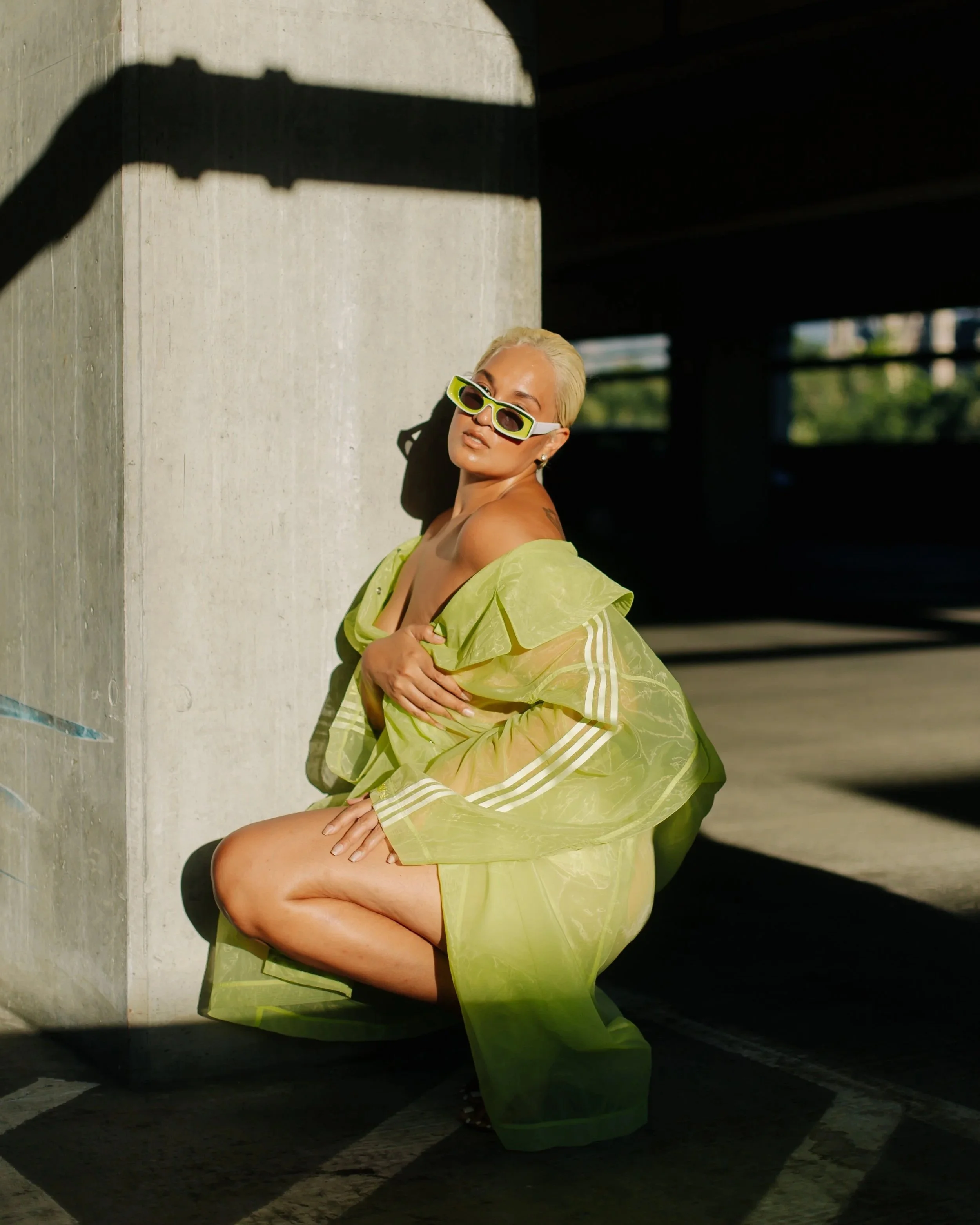 A woman with short, platinum blonde hair and sunglasses, wearing a sheer, lime-green dress, squatting next to a concrete pillar in an outdoor parking garage.