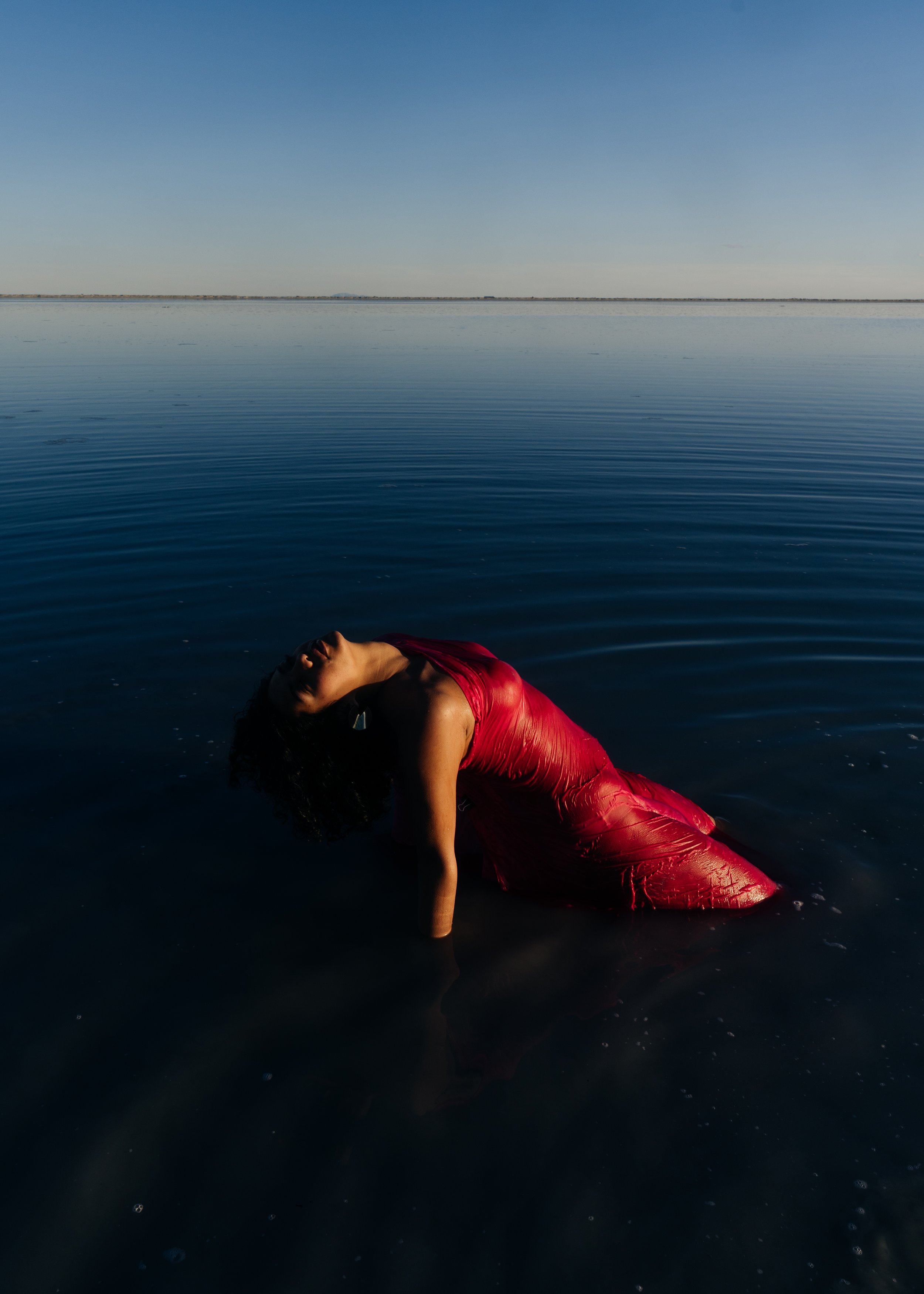 A woman in a red dress partially submerged in dark water, with her head tilted back and eyes closed, during sunset or sunrise on calm waters.