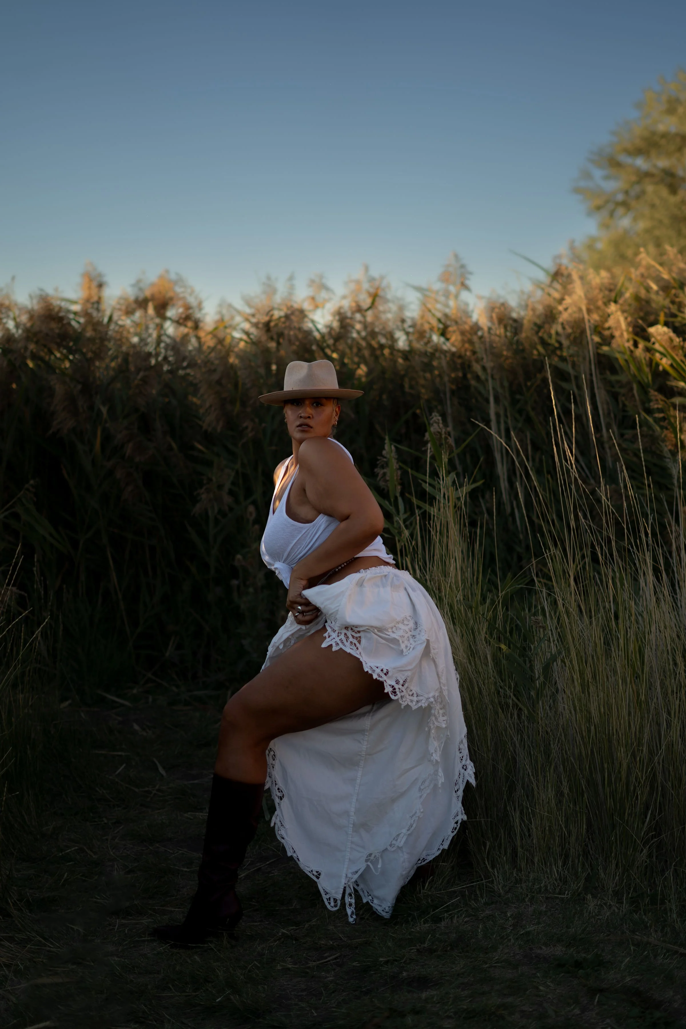 A woman wearing a white dress, a beige hat, and black boots standing in tall grass with a background of bushes and a blue sky.
