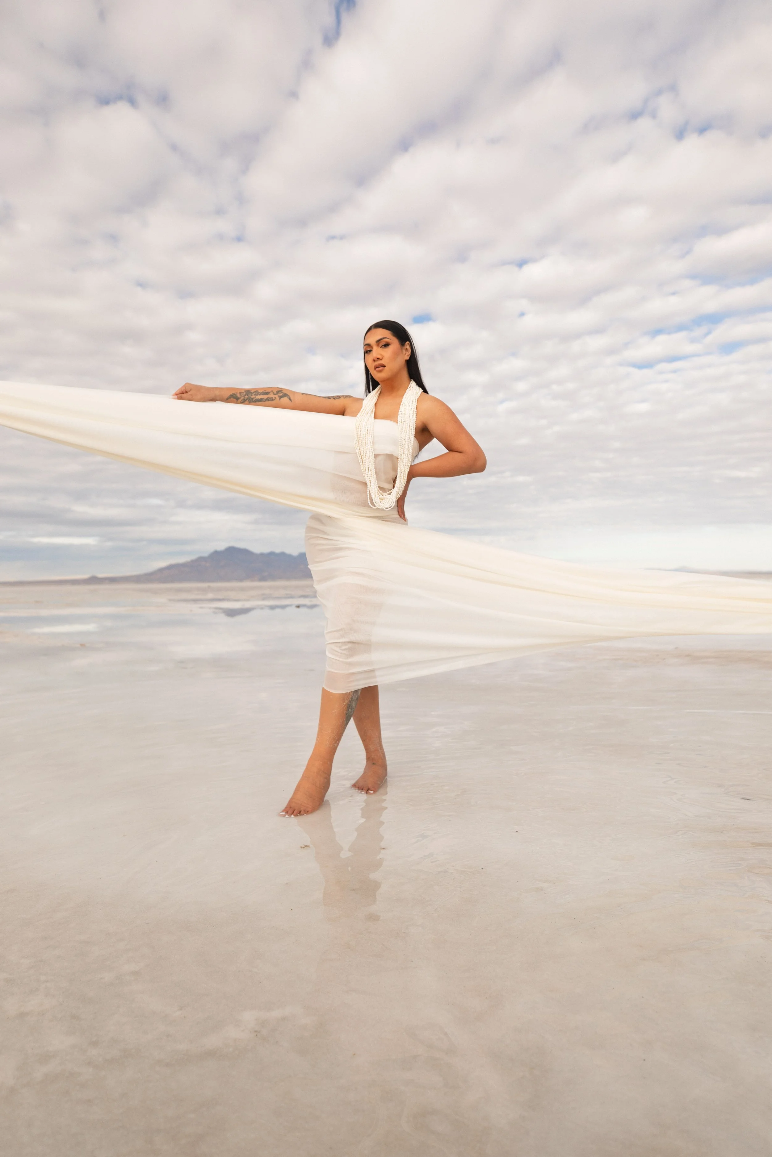 A woman in a white dress and multiple pearl necklaces posing on a reflective, sandy surface with mountains in the distance and a cloudy sky overhead.