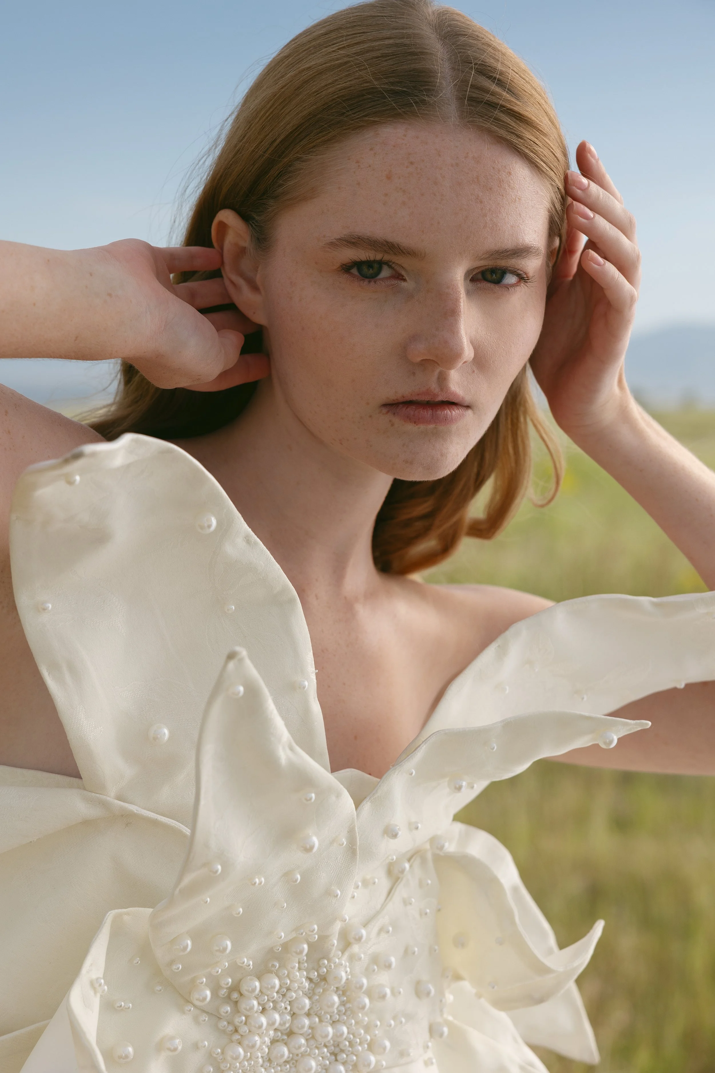 A young woman with red hair and freckles is outdoors, wearing a white dress with pearl embellishments, touching her ears, with a blurred natural background.