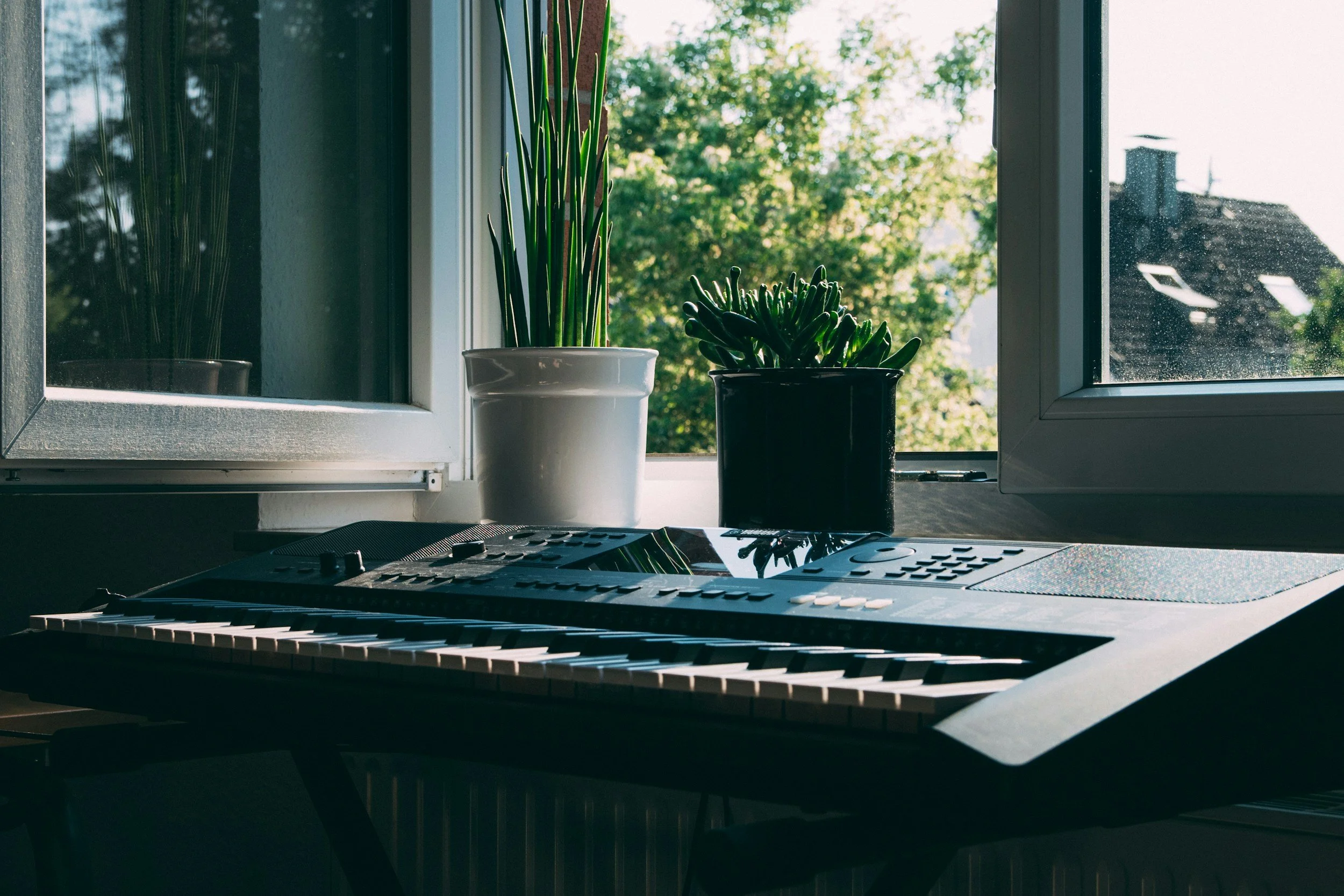 A digital keyboard on a desk in front of a window with two potted plants and a view of trees and rooftops outside.