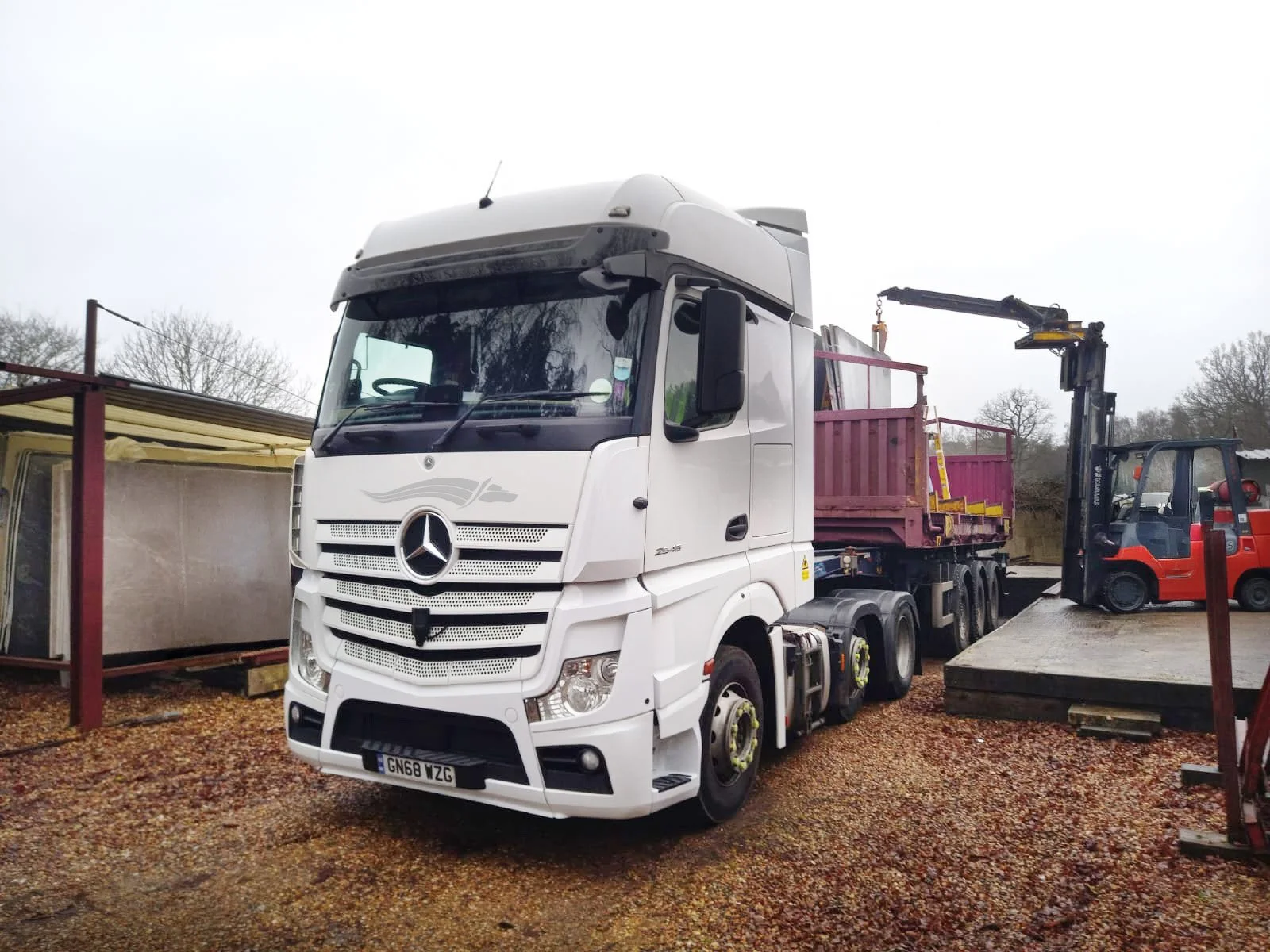 A white Mercedes-Benz semi-truck with a purple flatbed trailer parked outdoors on gravel, with a red forklift nearby lifting an item, and other outdoor equipment and structures in the background.
