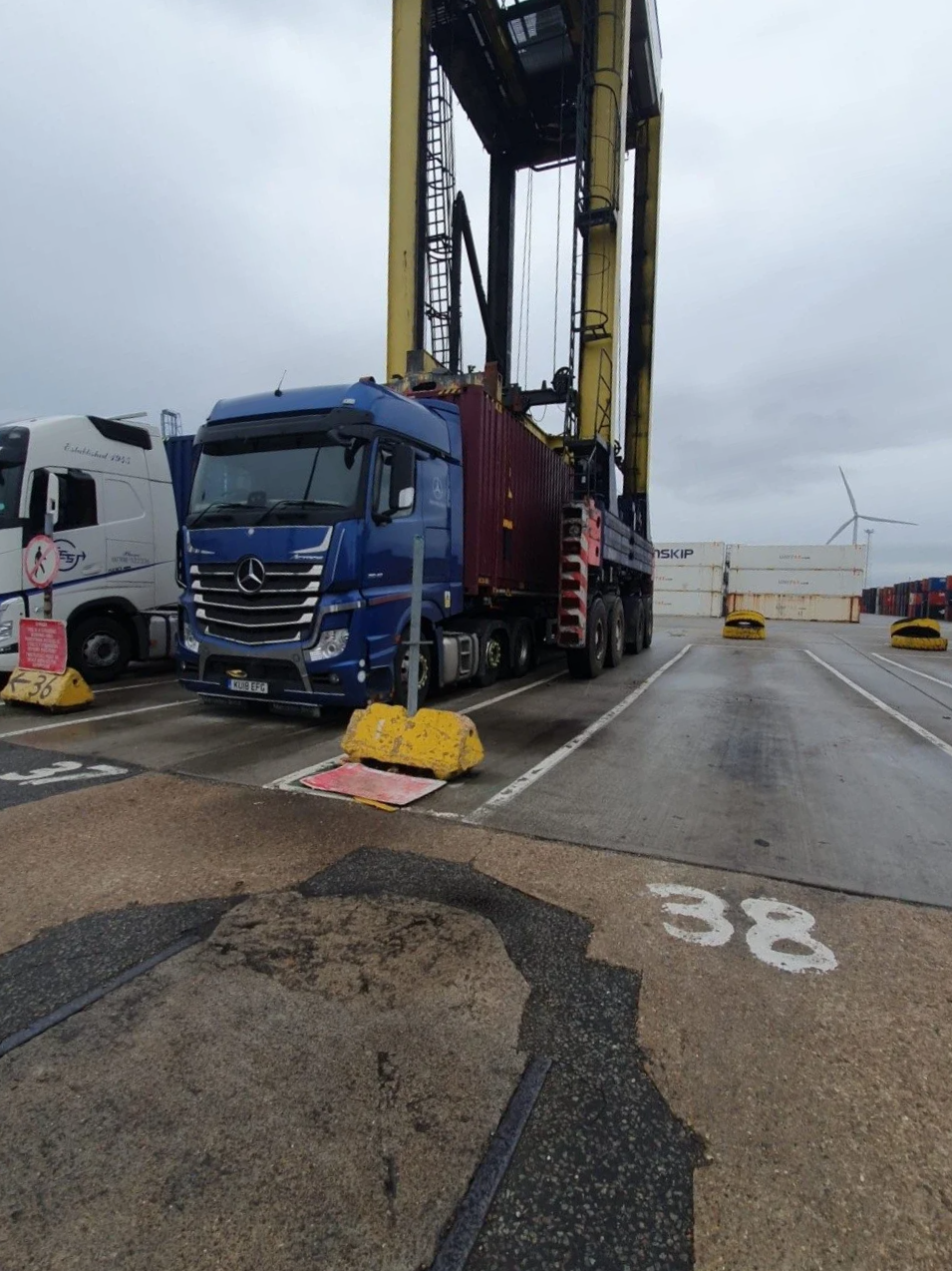 A blue truck with a large yellow crane lifting container at a port, with surrounding parked trucks and shipping containers in the background, and a cloudy sky overhead.