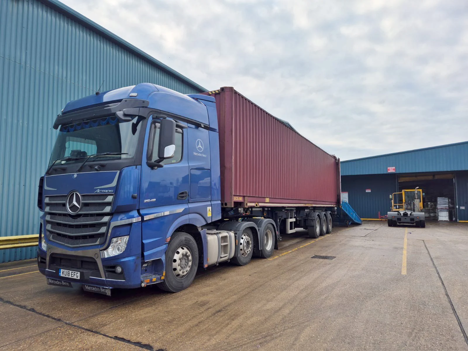 A large blue Mercedes-Benz semi-truck with a red cargo container parked at a loading dock in an industrial area, with a forklift nearby.