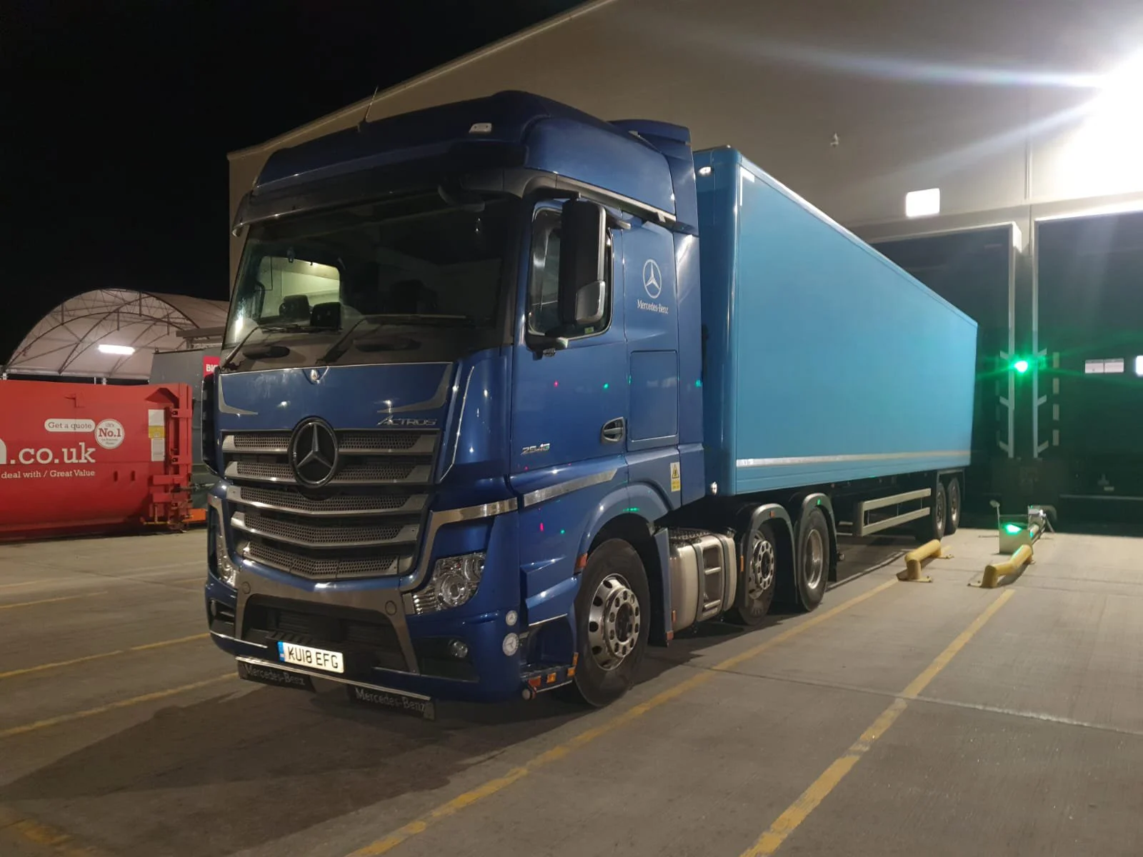A blue Mercedes-Benz truck parked in a parking lot at night, illuminated by nearby lights, with a red container on the left and a green light in the background.