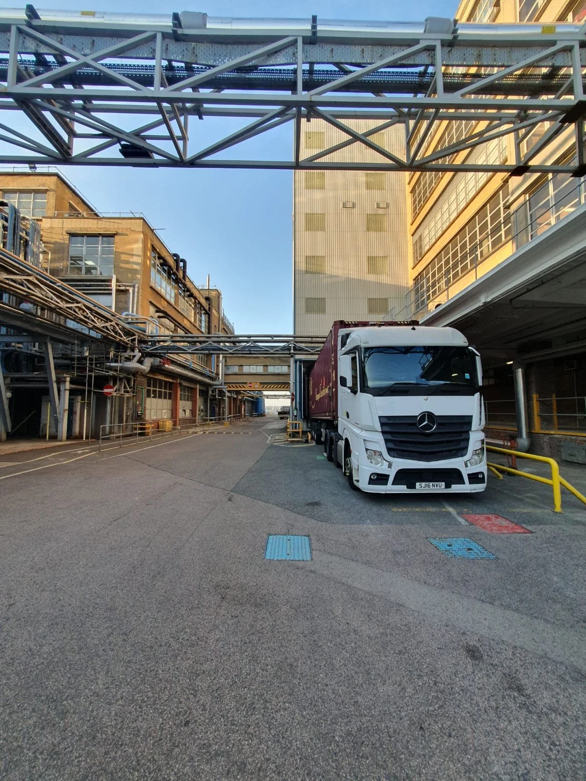 A white Mercedes-Benz semi-truck with a maroon trailer parked in a city industrial area under a metal bridge, with tall buildings in the background and a blue sky.