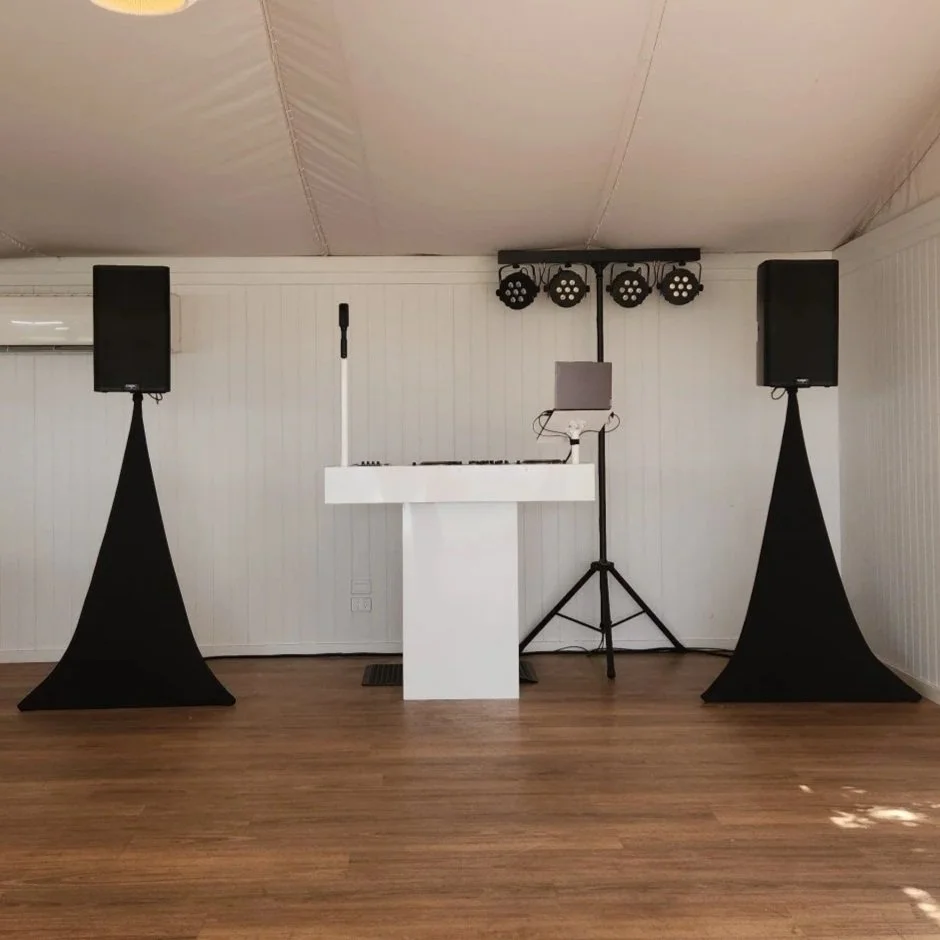 DJ setup with two large speakers on black stands, a DJ controller on a white table, and stage lighting on a wall in a room with wood flooring and white paneled walls.