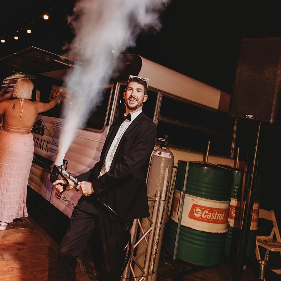 Man in tuxedo holding a flame effect device at a nighttime event, with a woman in a pink dress and food truck in the background.