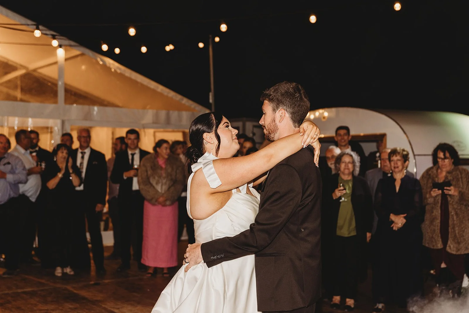 A bride and groom share their first dance at night, surrounded by wedding guests under string lights.