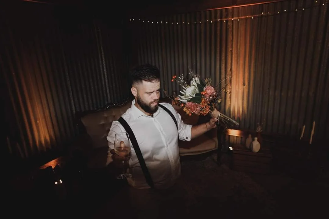 A man with a beard and white shirt holding a glass of wine in one hand and a bouquet of flowers in the other, standing in a dimly lit room with wooden walls.