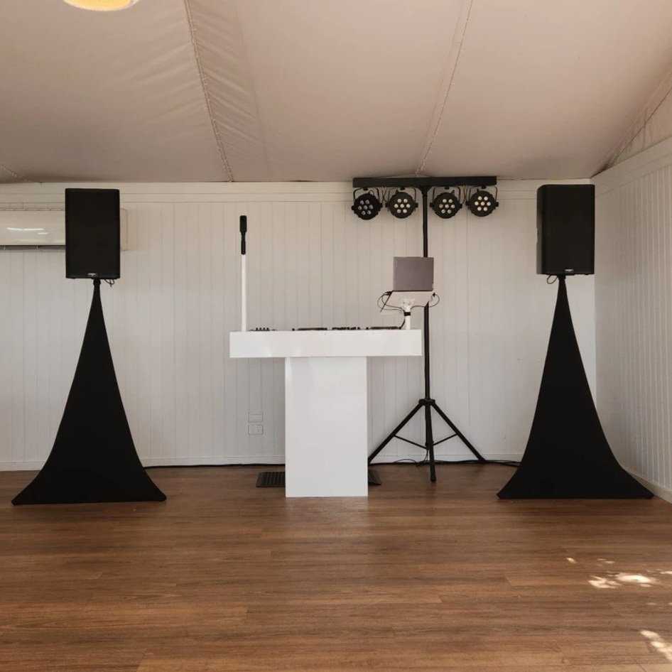 DJ setup with speakers, a microphone on a stand, and stage lighting in a white-walled room with wooden floor.