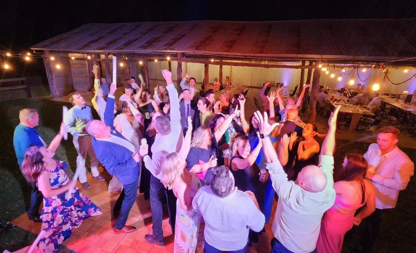 Guests dancing and celebrating at an outdoor nighttime party with string lights, some holding drinks, under a rustic wooden shelter.
