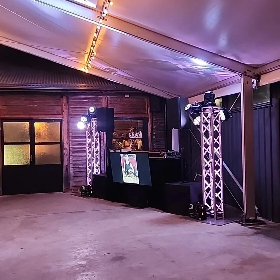 Event space with stage, DJ booth, and lighting equipment under a white tent, featuring wooden walls and frosted windows.