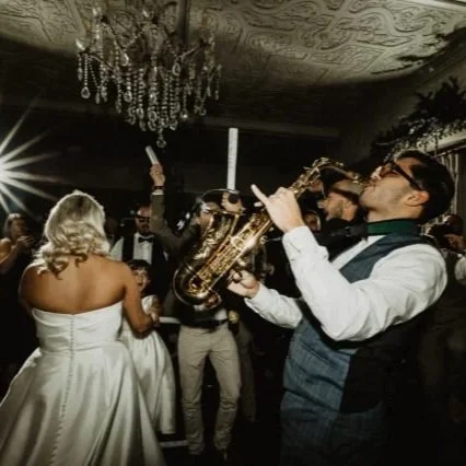 Man playing saxophone at a wedding reception with a bride and other guests in a dimly lit room with a chandelier.