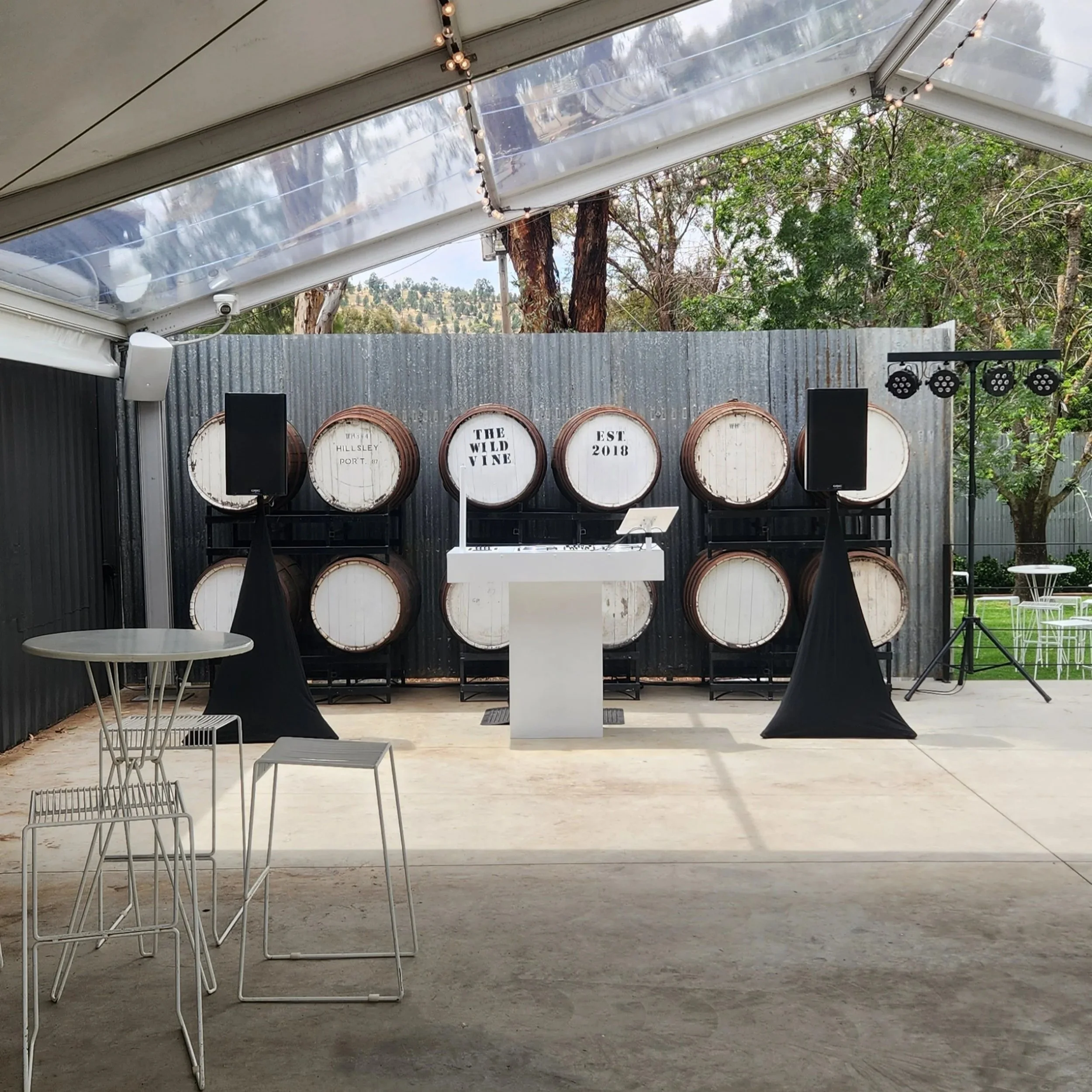 Wedding reception setup with wine barrels, speakers, a DJ booth, string lights, and outdoor tables and chairs.