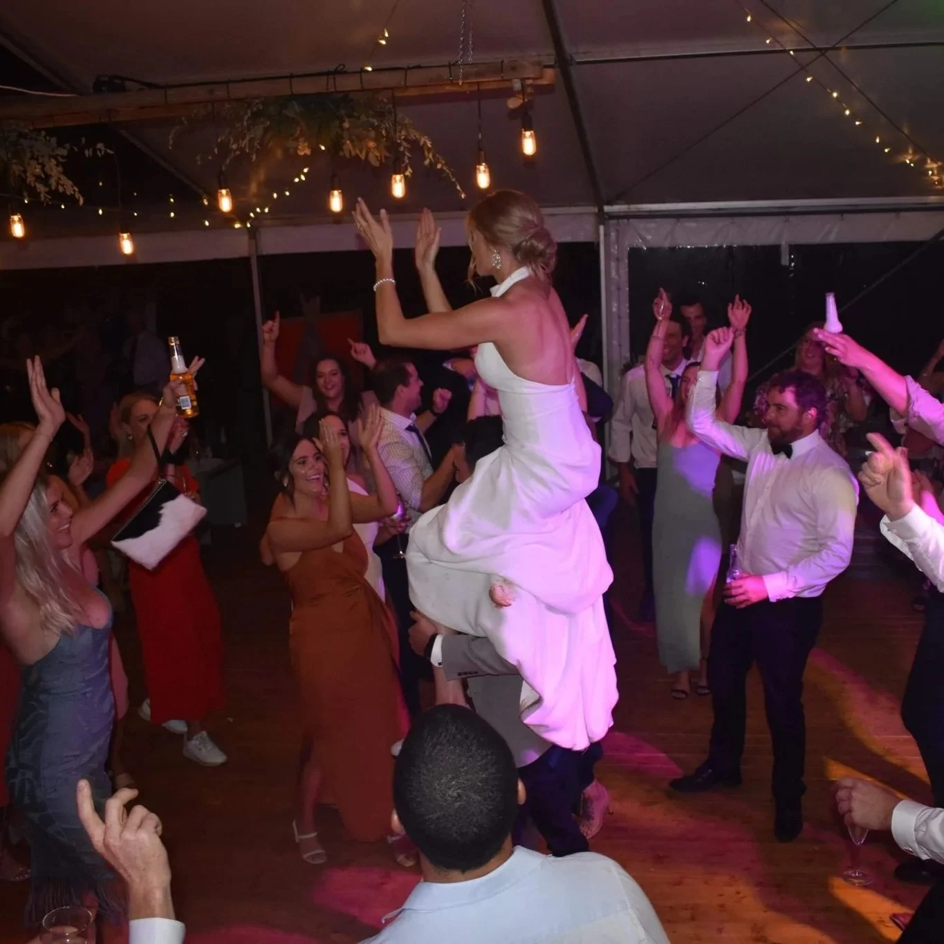 A bride in a white wedding dress is being lifted on a person's shoulders during a wedding reception, dancing and celebrating with guests under warm string lights.