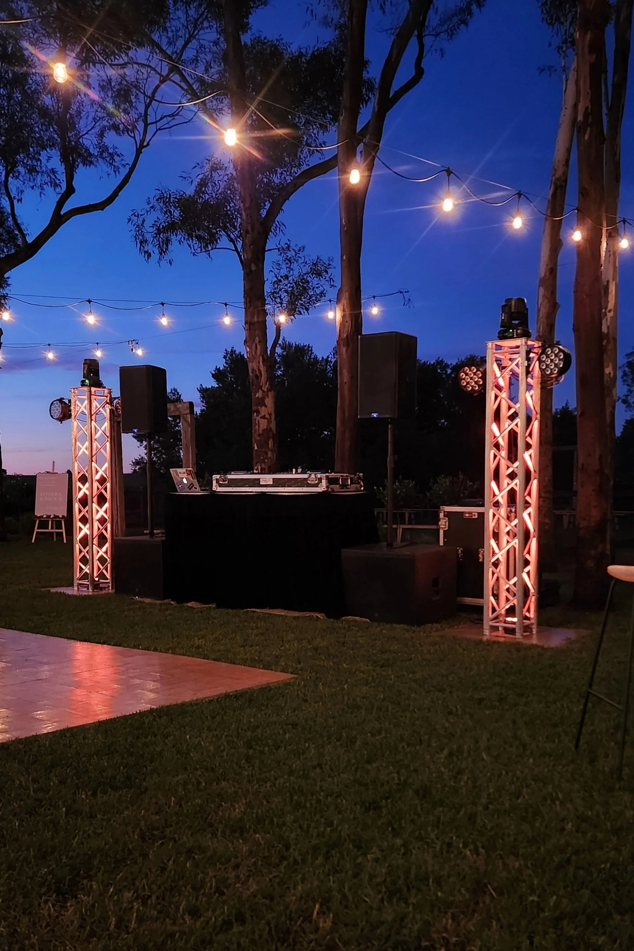 Outdoor evening setting with string lights, tall trees, a DJ booth with speakers, and lighting equipment on stage, suggesting event or dance party