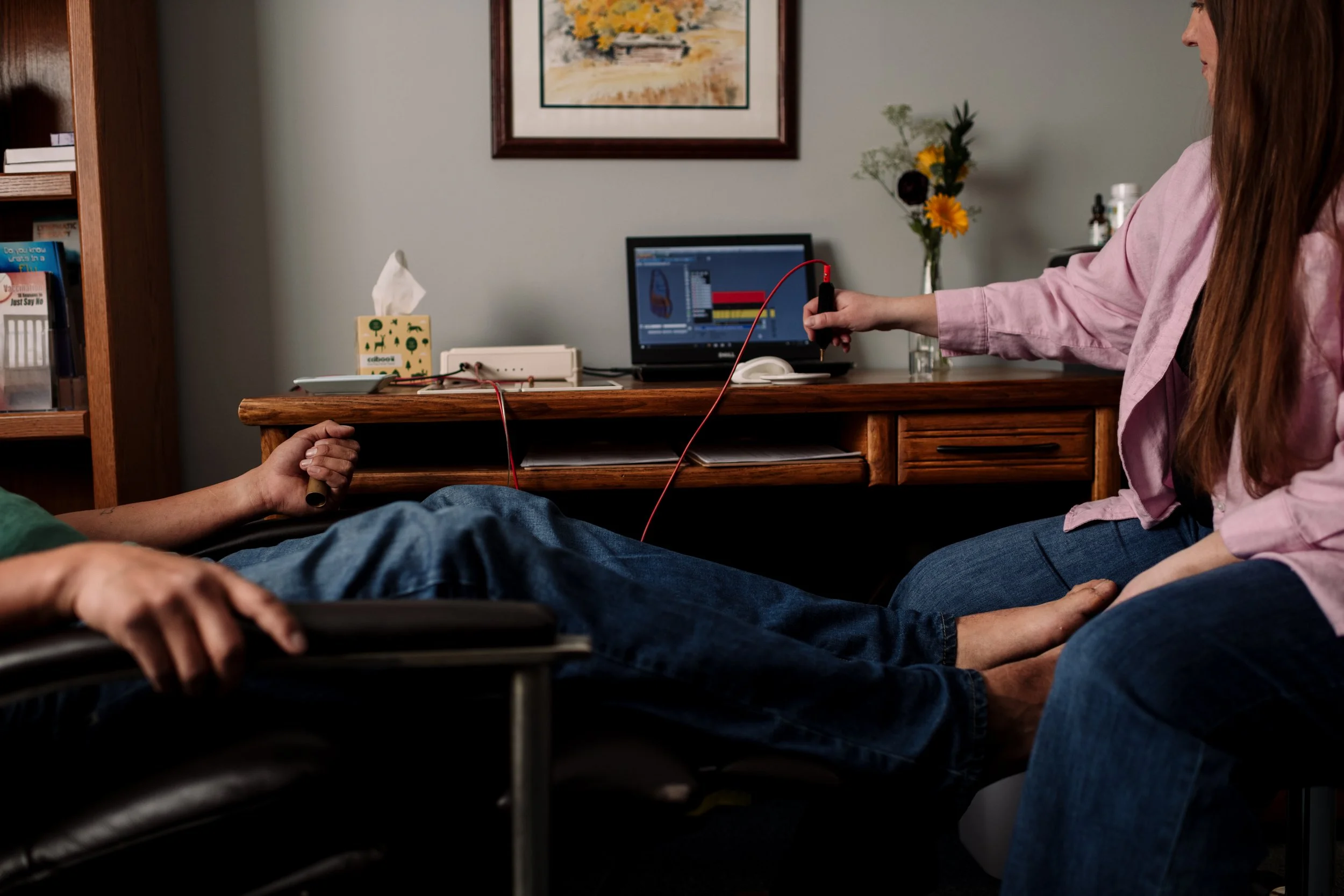 A person lying in a wheelchair connected to a monitoring device, with another person sitting beside them holding a probe, in a room with a desk, a computer, and a framed painting on the wall.