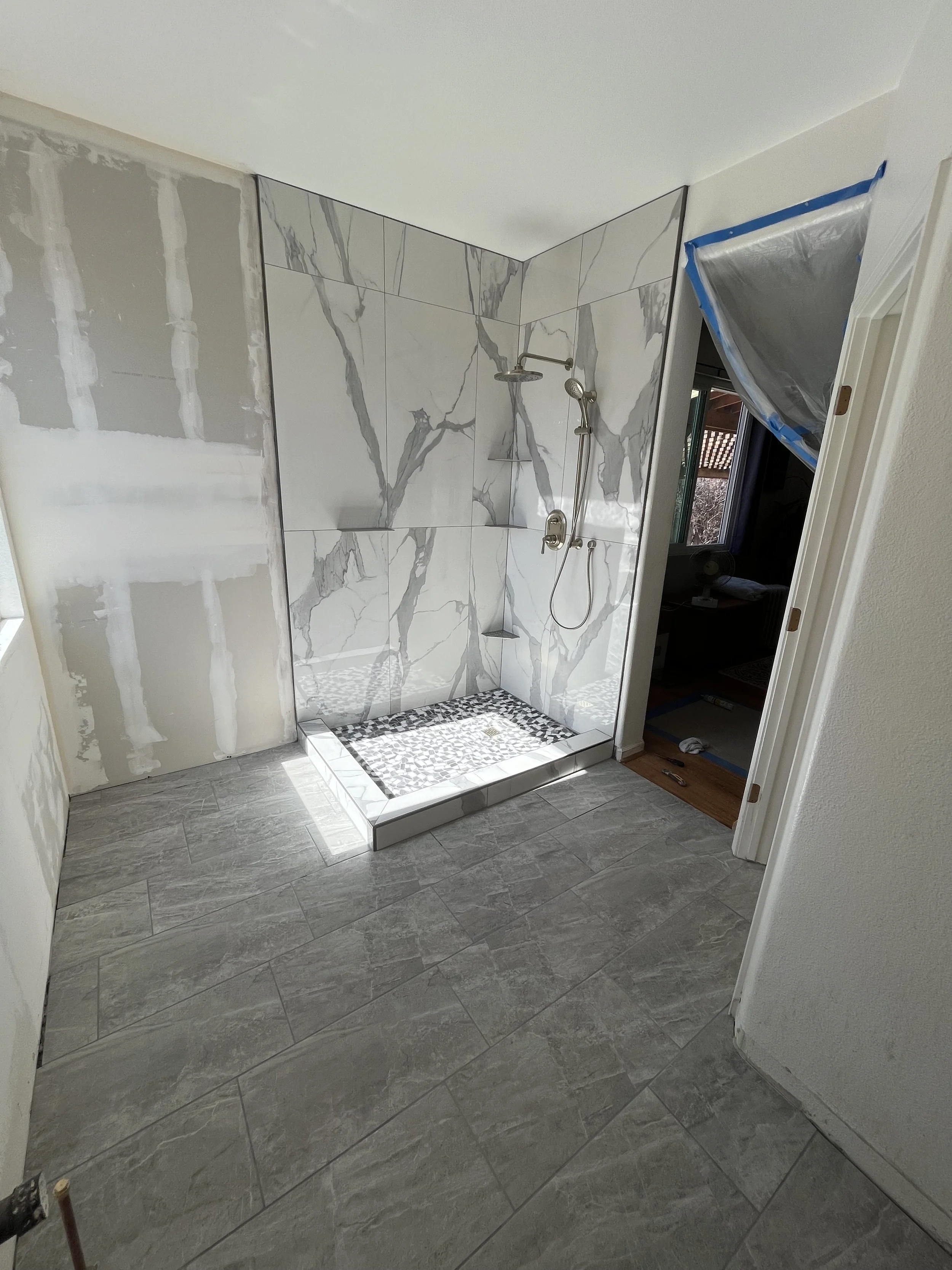 A bathroom with a tiled walk-in shower featuring large white marble tiles with gray veining, a rain showerhead, and a handheld showerhead. The shower base has small mosaic tiles. The bathroom walls are partially unfinished, with drywall and joint compound visible, and a doorway with a plastic covering on the door frame is on the right.
