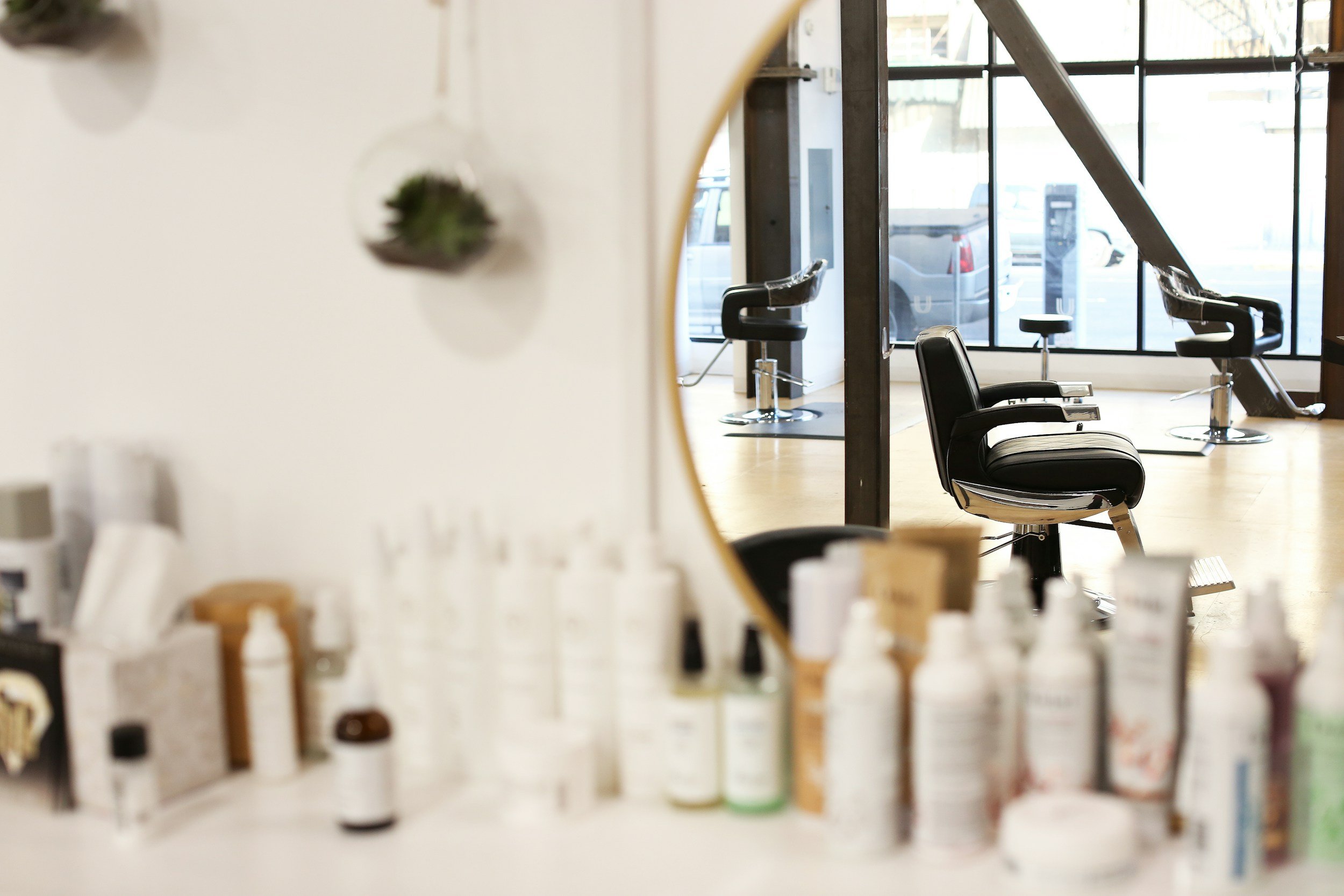Empty black salon chairs in a hair salon with large windows and wooden flooring, viewed through a mirror and blurred hair products in the foreground.