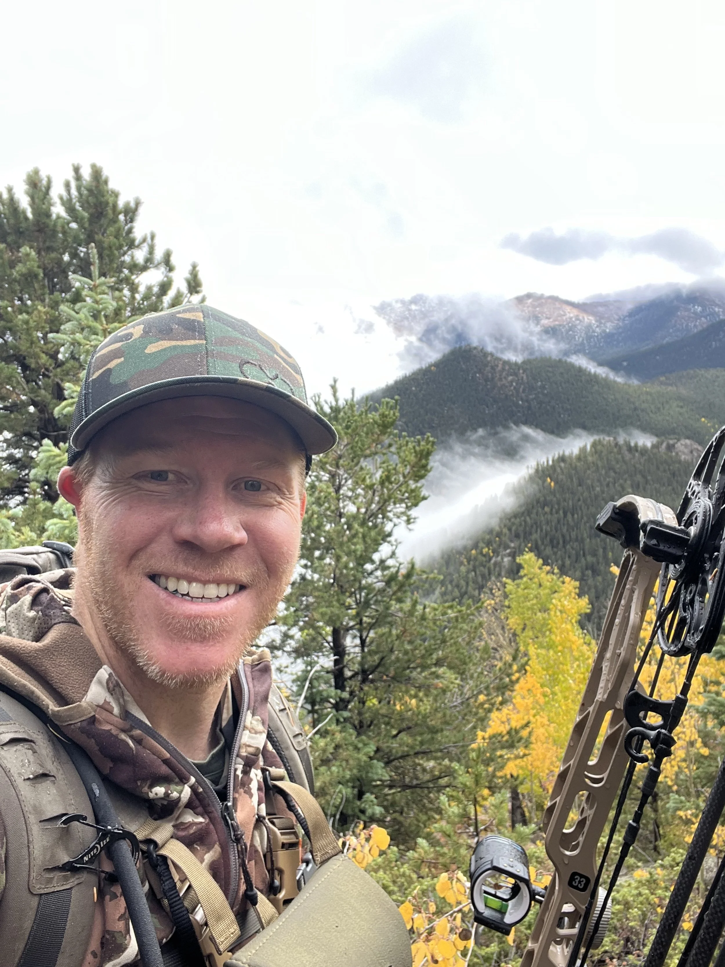 A man smiling outdoors in a mountain forest with fall foliage, holding a bow, with mountains and clouds in the background.