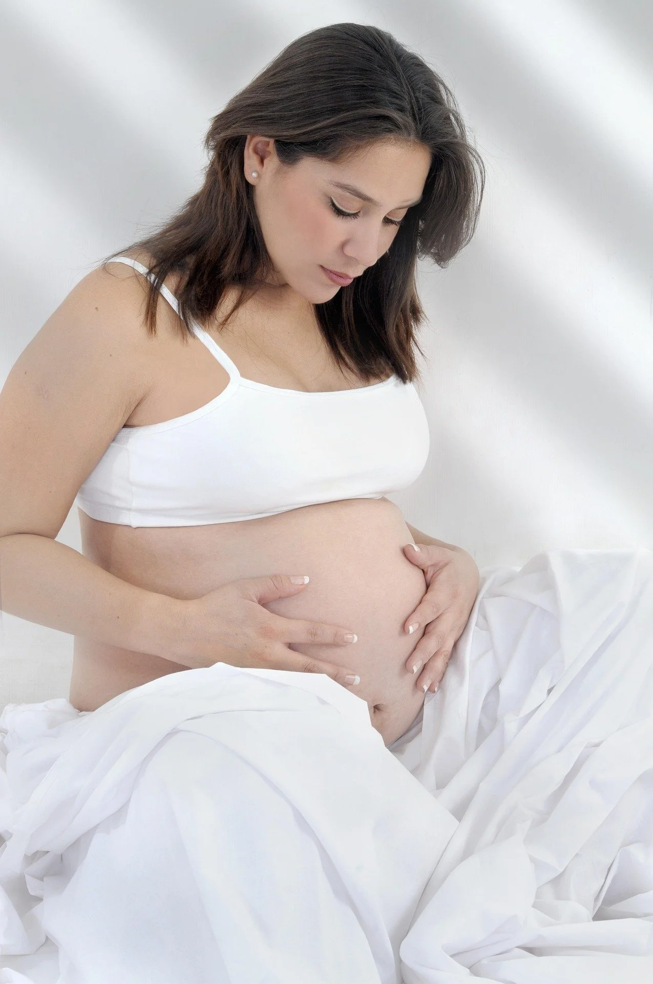 A pregnant woman in a white top gently cradles her exposed belly with her hands, sitting on a white bed with a soft, blurred background.