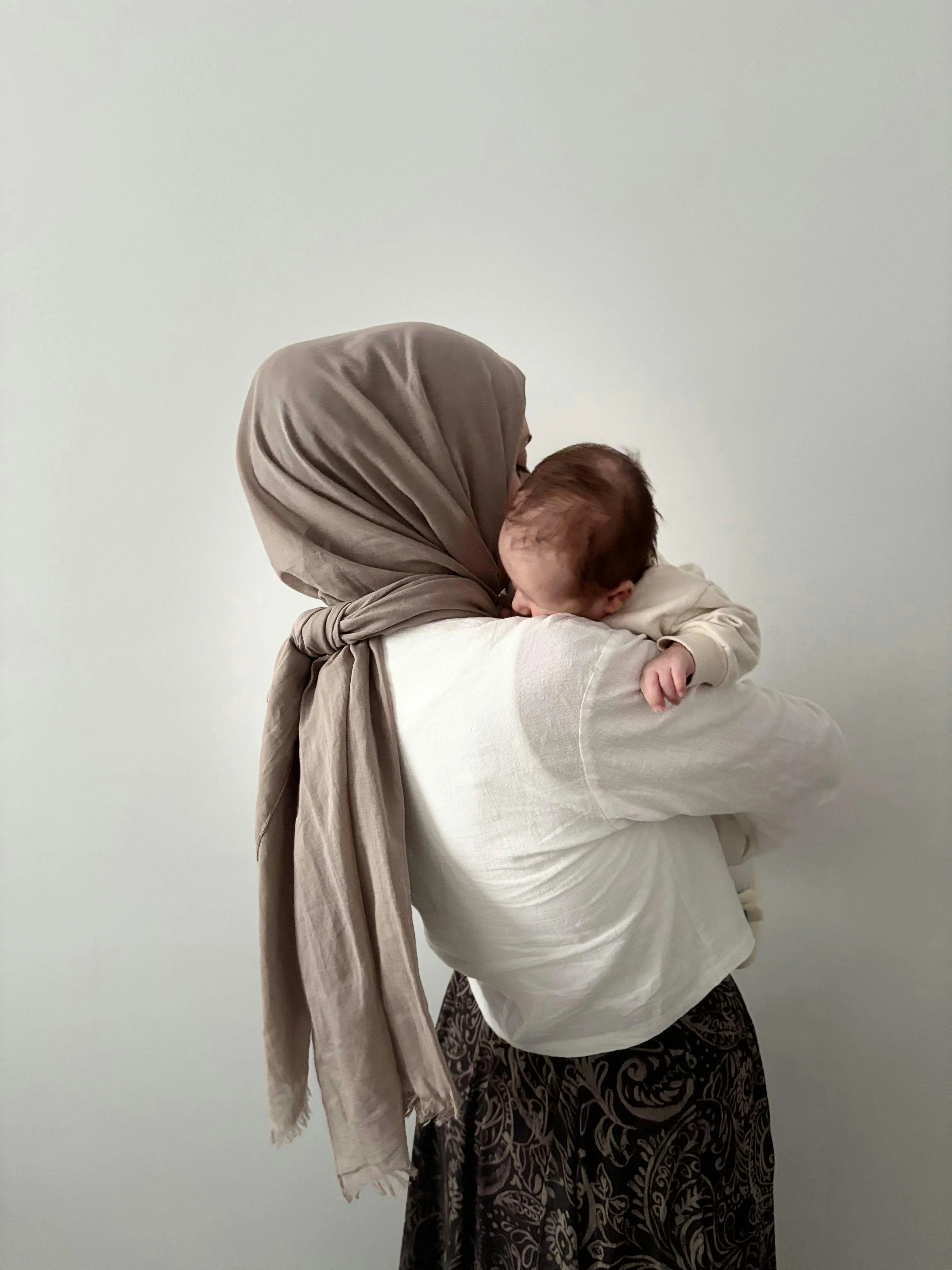 A woman in a beige headscarf holding a sleeping baby against a plain white wall.