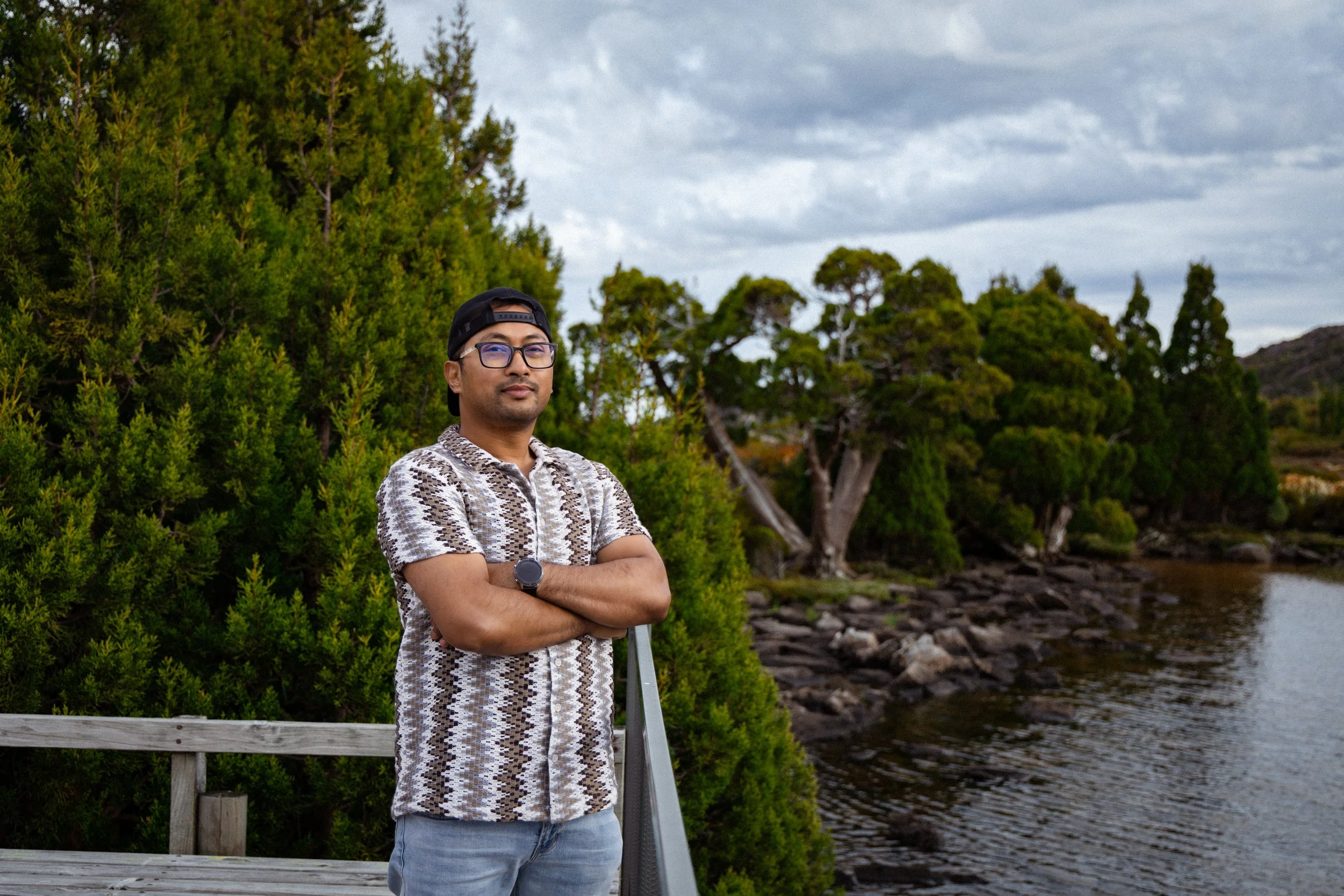 A man standing on a wooden bridge with arms crossed, wearing glasses, a patterned shirt, a black cap worn backwards, and a watch, near a river with lush green trees and a cloudy sky in the background.
