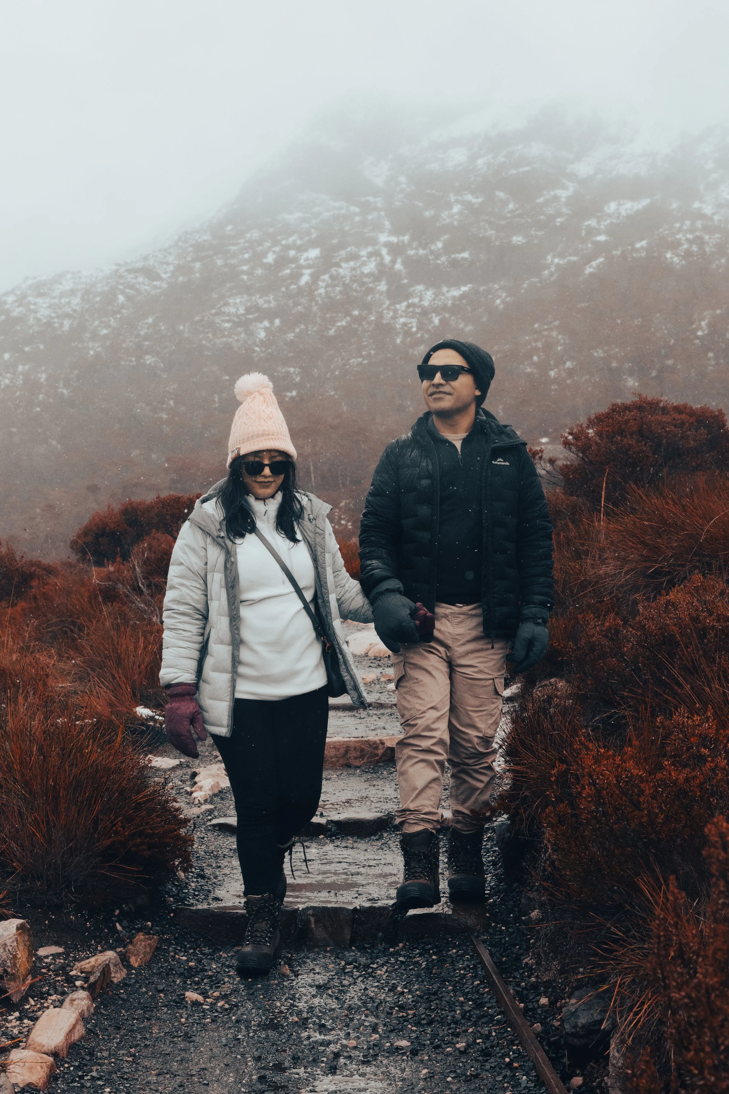 A man and woman hiking on a rocky trail with orange bushes and foggy mountains in the background.