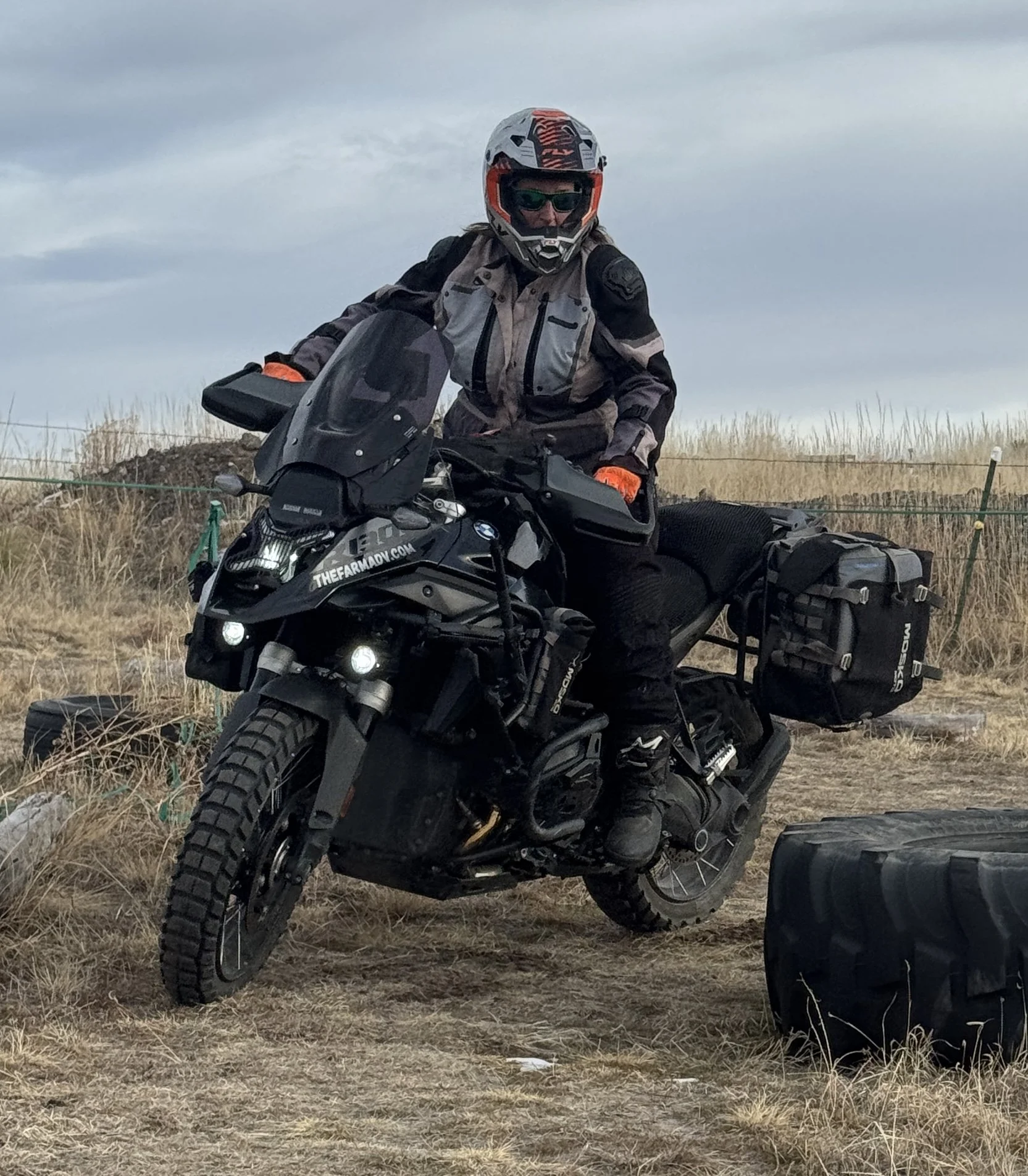 Person wearing a helmet and sunglasses riding a black adventure motorcycle in a grassy field with a cloudy sky in the background.