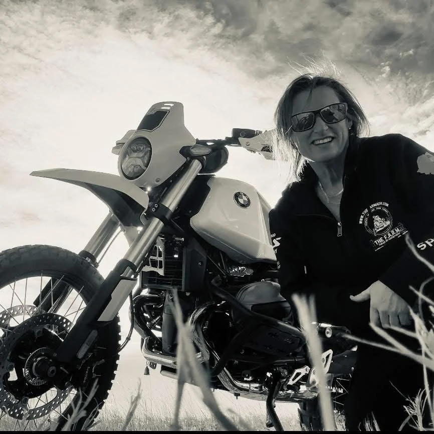 A woman smiling and wearing sunglasses, kneeling next to a BMW motorcycle in an outdoor grassy area, with cloudy sky in the background.