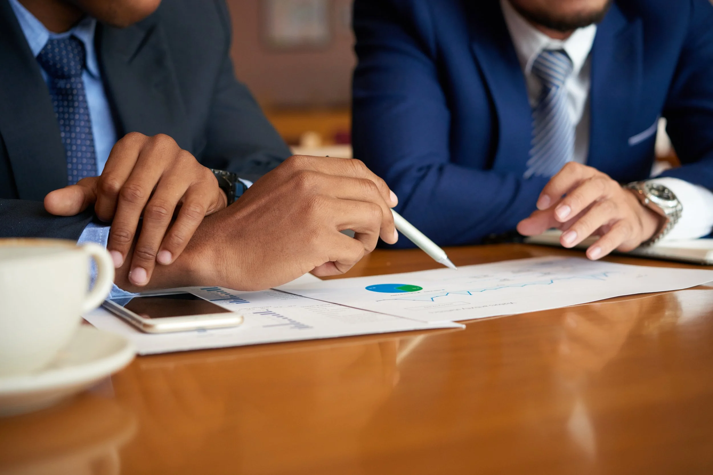 Two men in suits discussing financial graphs and documents at a wooden table, with a cup and a smartphone nearby.