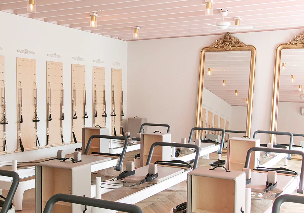 A Pilates studio with reformer machines arranged in rows, mirrors with ornate gold frames, and a light pink ceiling with recessed lighting.
