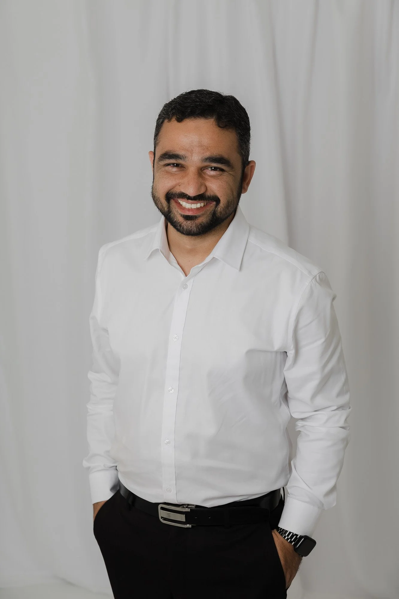 Man wearing a white dress shirt, black pants, black belt, and a black watch, smiling, standing against a plain background.