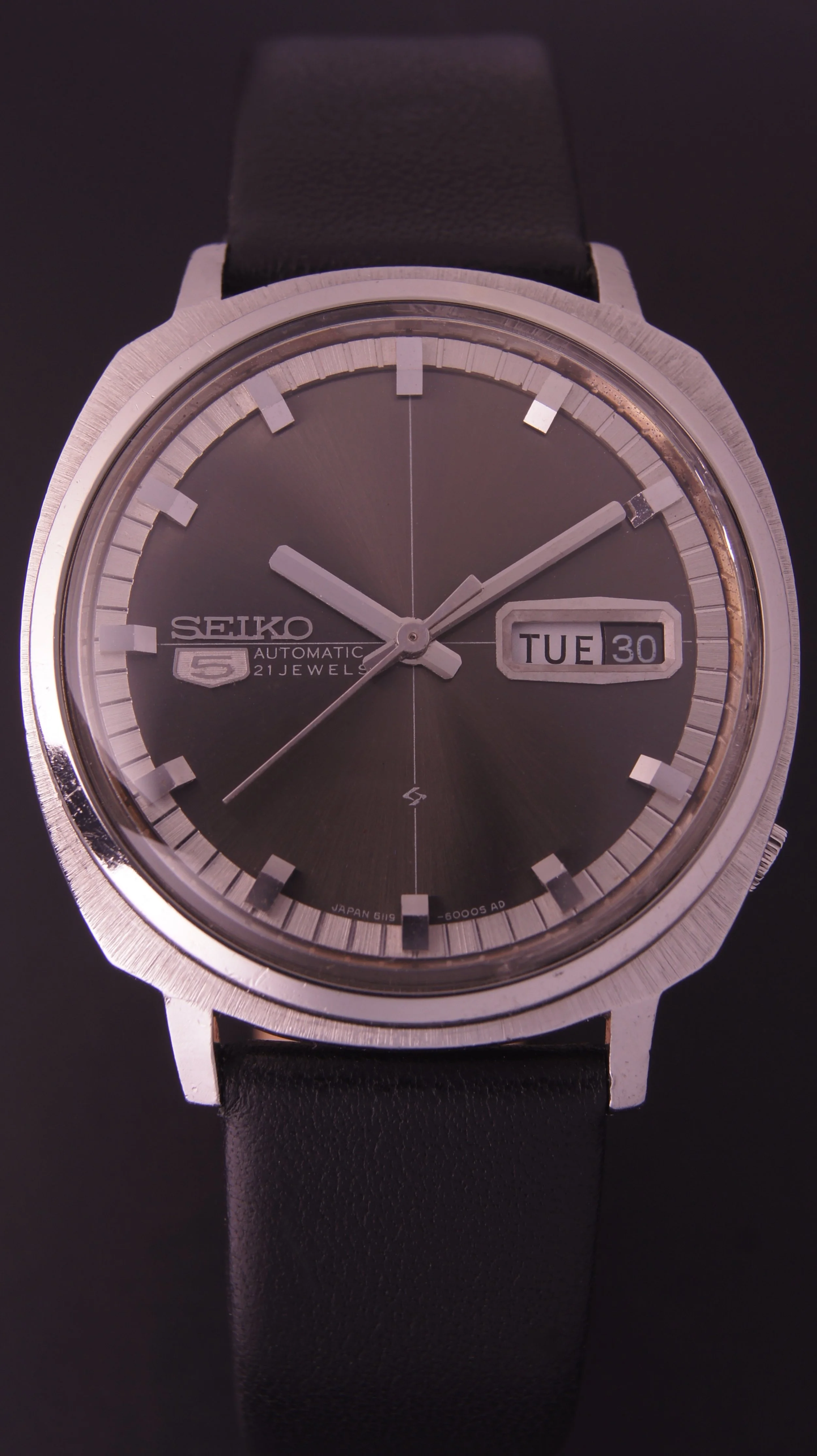 A Seiko automatic wristwatch with a black dial, silver hour markers, and a black leather strap, showing the time as 10:08 and the date as Tuesday the 30th.