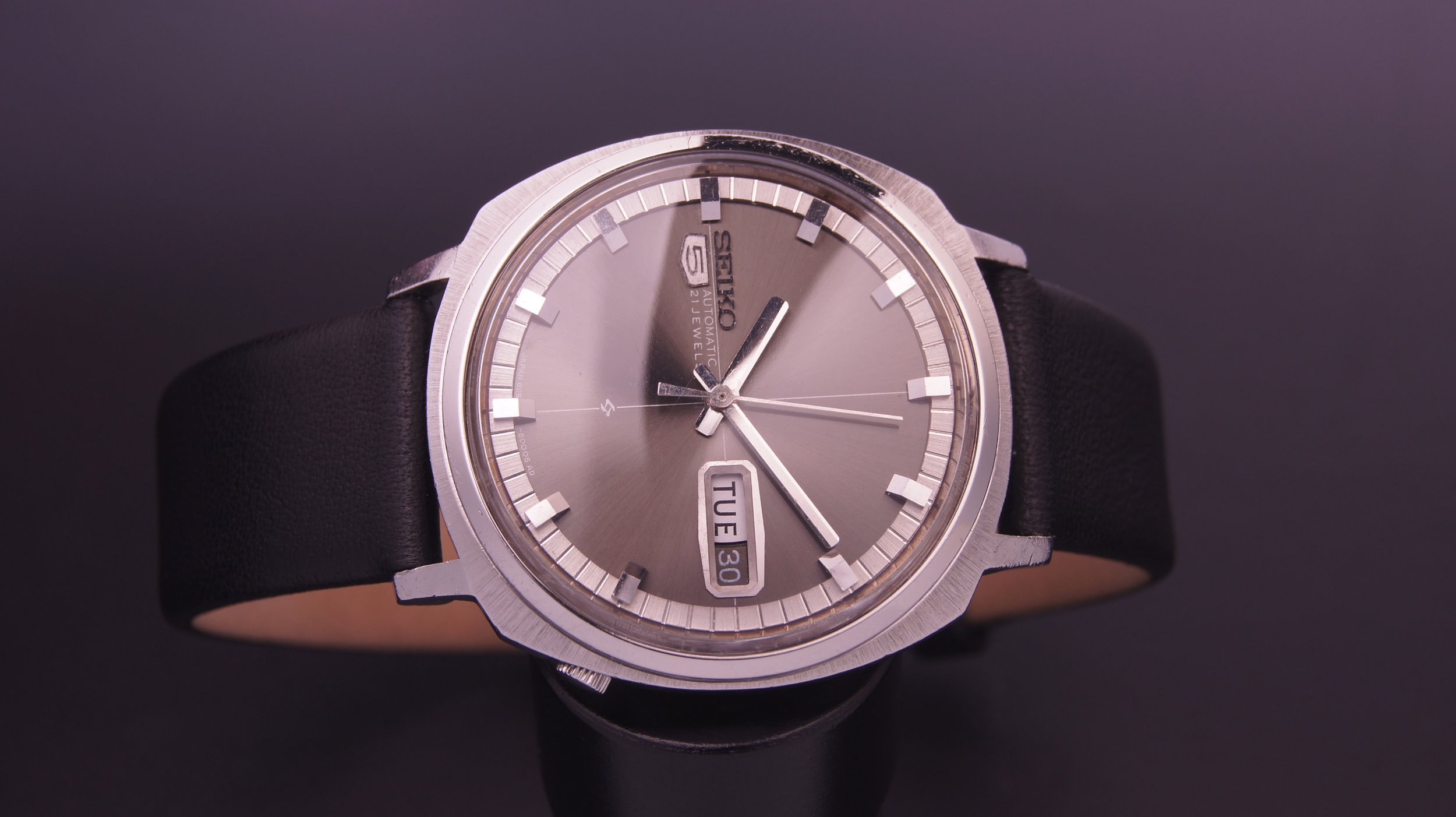 A vintage Seiko automatic wristwatch with a silver face, day and date display, black leather strap, and silver hour and minute hands.