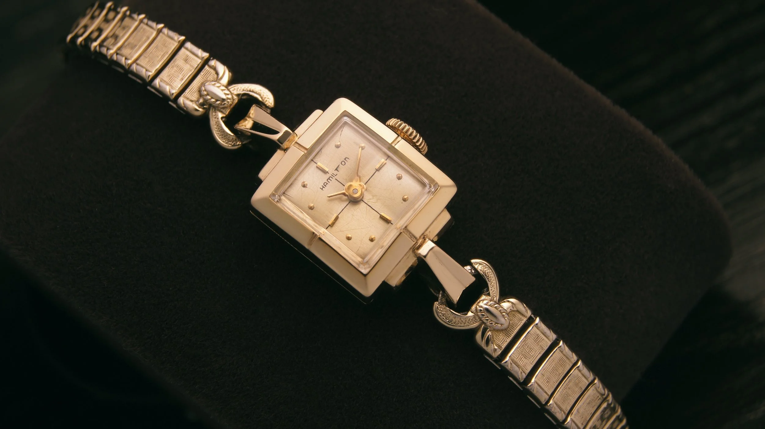 A vintage Hamilton women's wristwatch with a square gold face and a gold bracelet with decorative links, resting on a dark surface.
