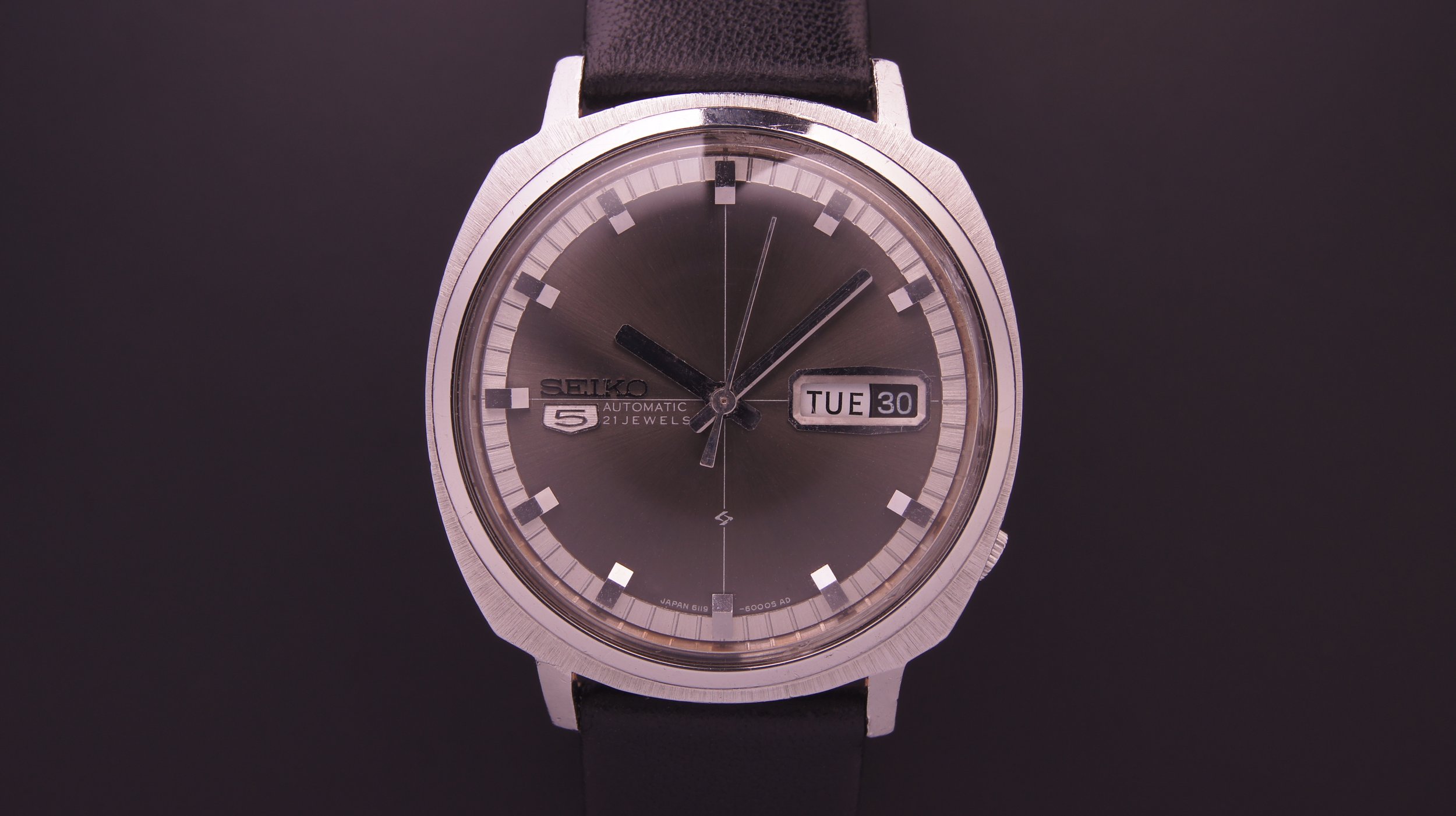 Close-up of a vintage Seiko automatic wristwatch with a black leather strap, dark gray dial, day and date display showing "TUE 30," and hour, minute, and second hands, set against a dark background.