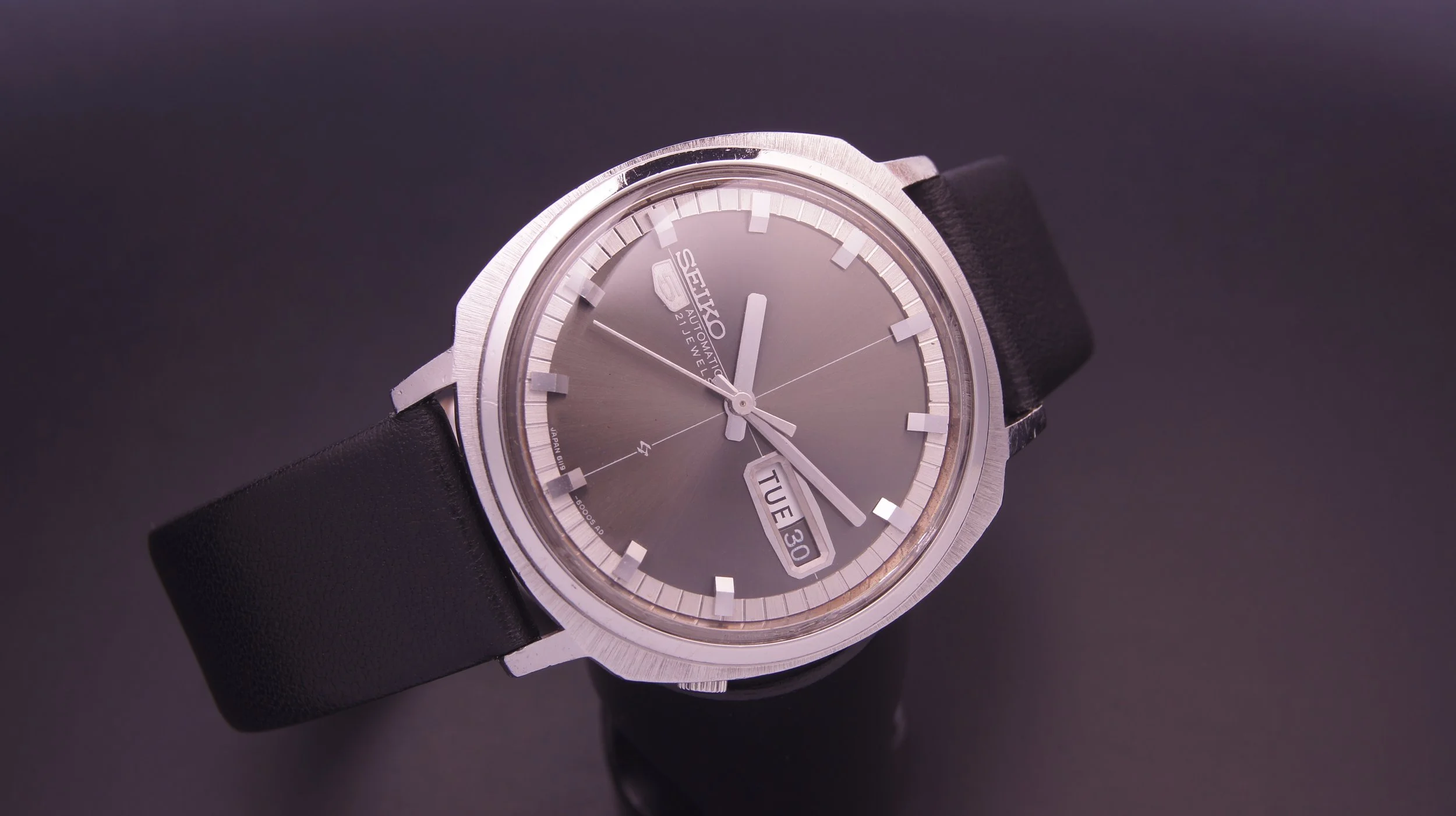 Close-up of a Seiko 5 automatic wristwatch with a gray dial, day and date display, black leather strap, and silver case on a dark background.