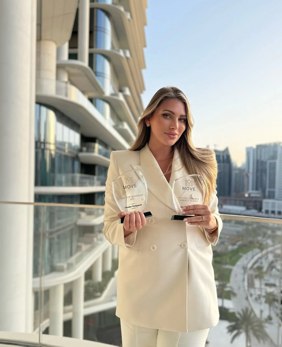 A woman in a cream-colored suit holding two awards on a balcony with a cityscape in the background.