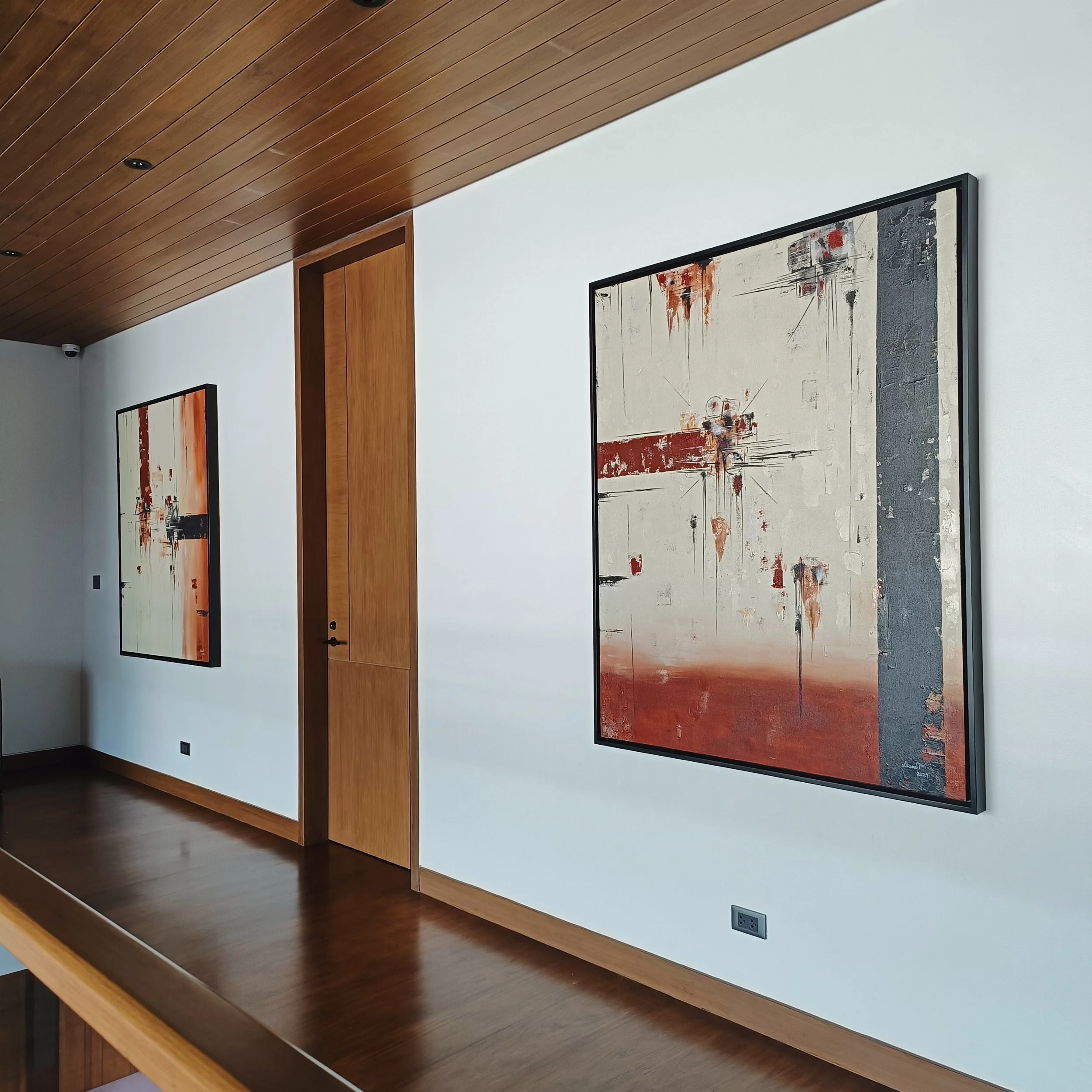 Interior view of a modern art gallery with two large abstract paintings on a white wall, featuring red, black, white, and gray tones, with a wooden ceiling and floor.