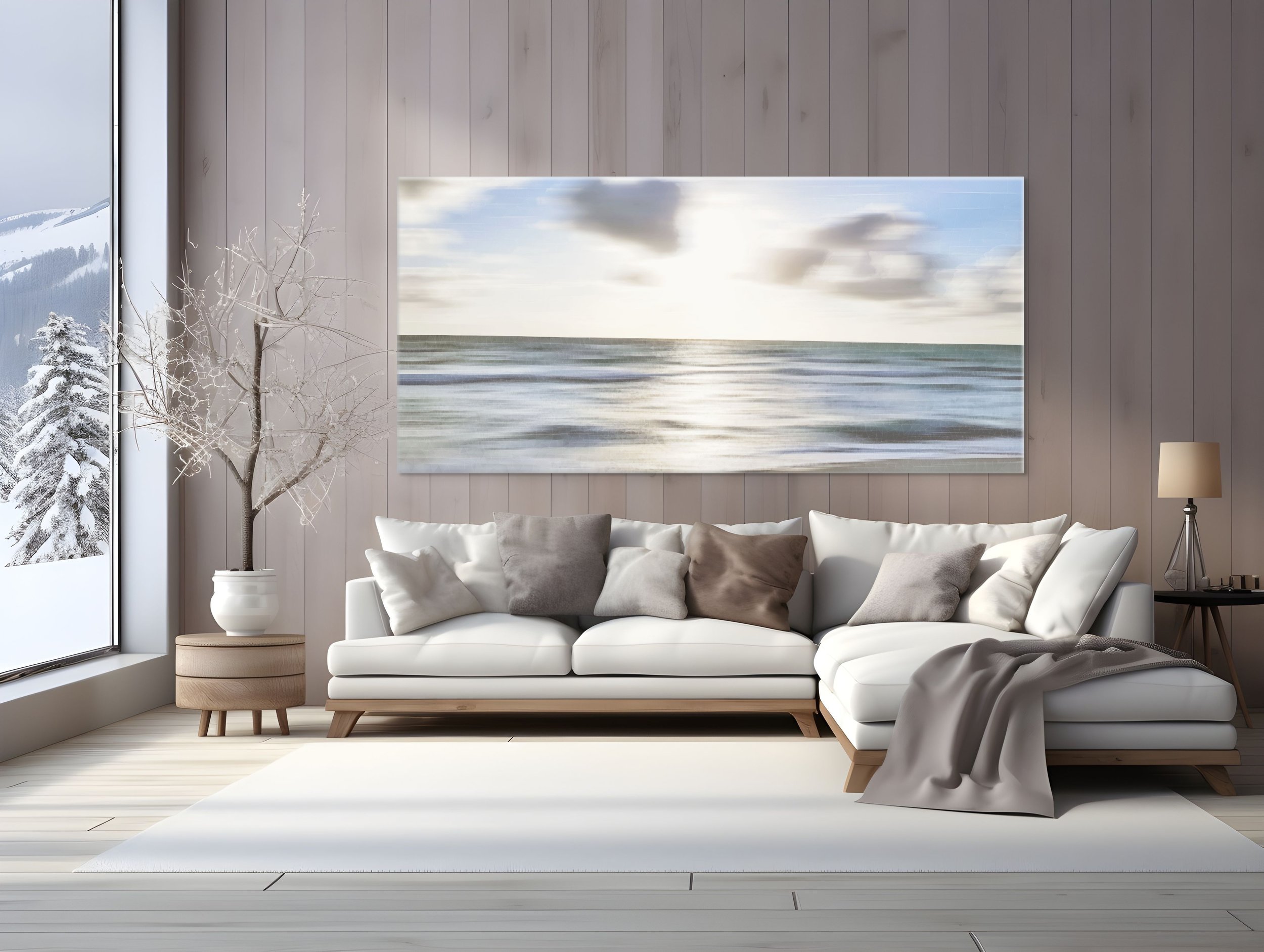 Modern living room with beige sofa, pillows, a side table with a lamp, large art piece of a seascape on the wall, and large windows showing snow-covered trees outside.