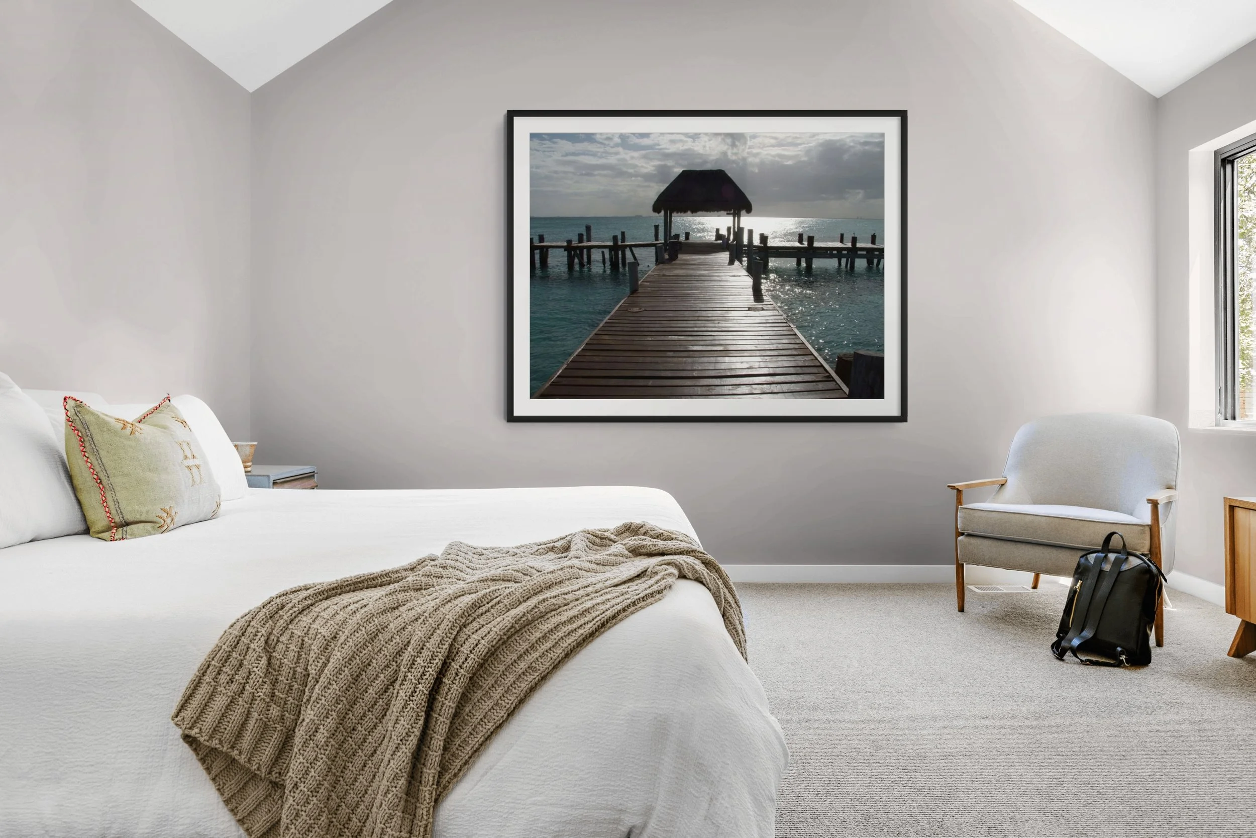 A bedroom with a large bed, a pillow with decorative embroidery, a blanket, and a framed picture of a pier extending into the ocean on a cloudy day. There is a white armchair, a small table, a backpack on the floor, and a window with a view outside.