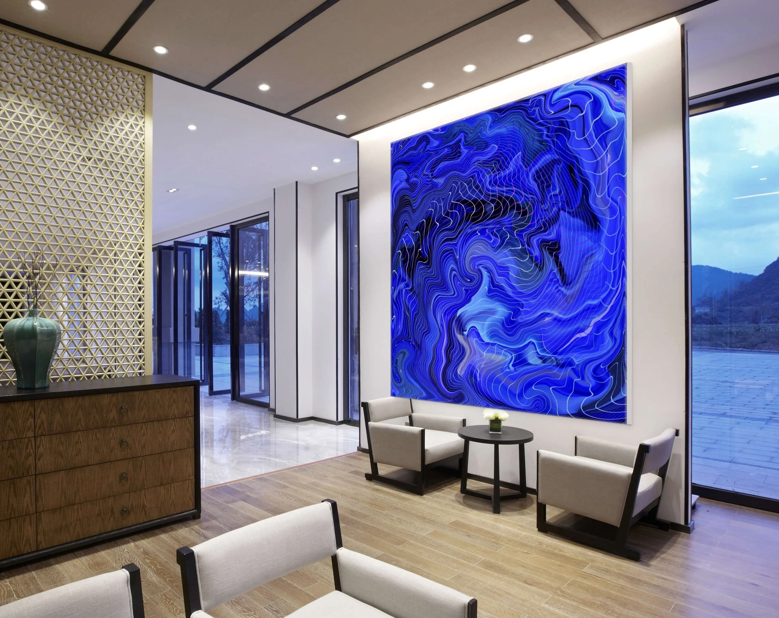 Modern hotel lobby with a large abstract blue artwork, seating area with beige chairs and black tables, wooden sideboard with a green vase, sliding glass doors, and windows showing mountain scenery.