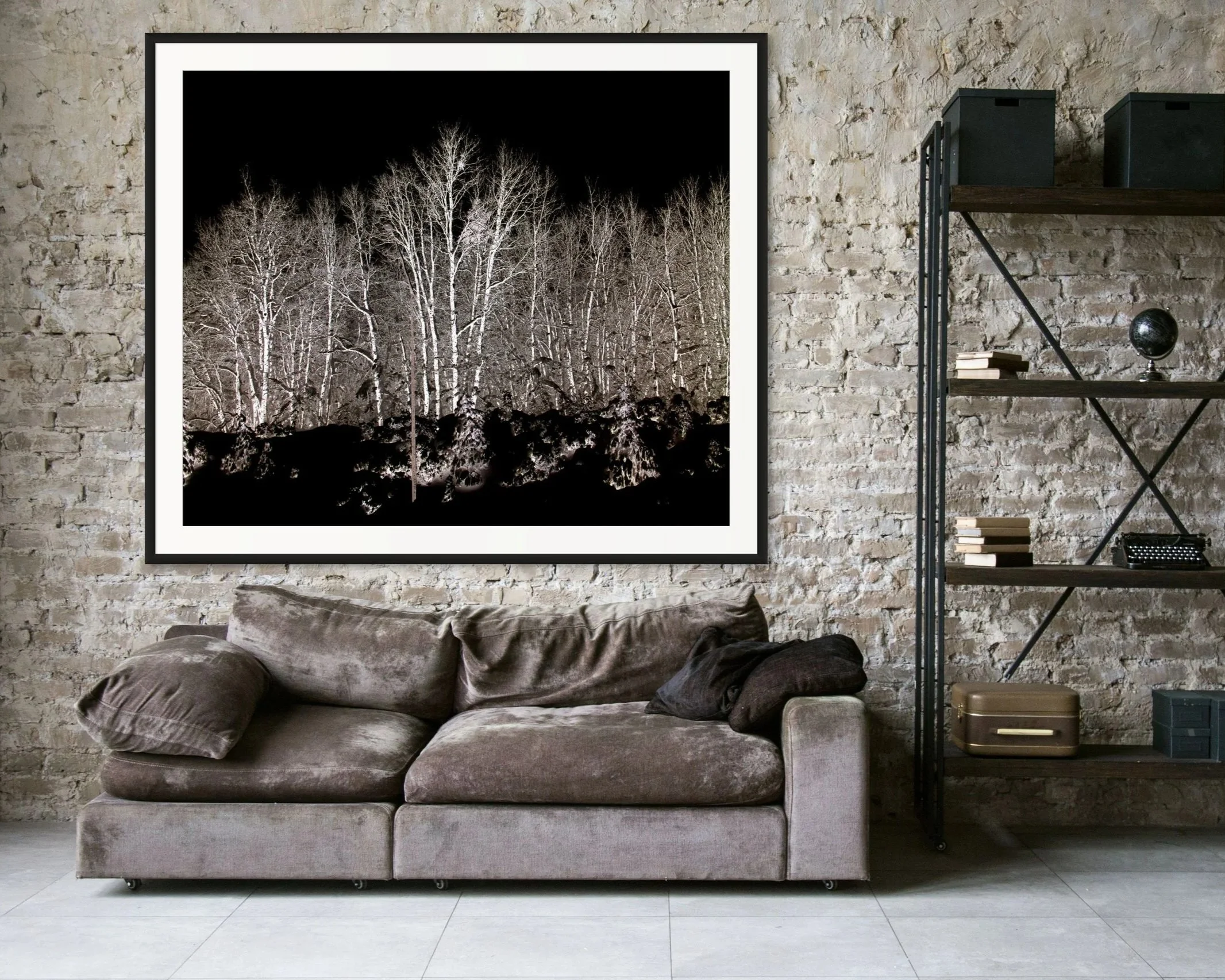 Living room with a textured brick wall, a framed black and white photo of leafless trees, a gray velvet sofa with pillows, and a bookshelf with books, a small globe, and storage boxes.