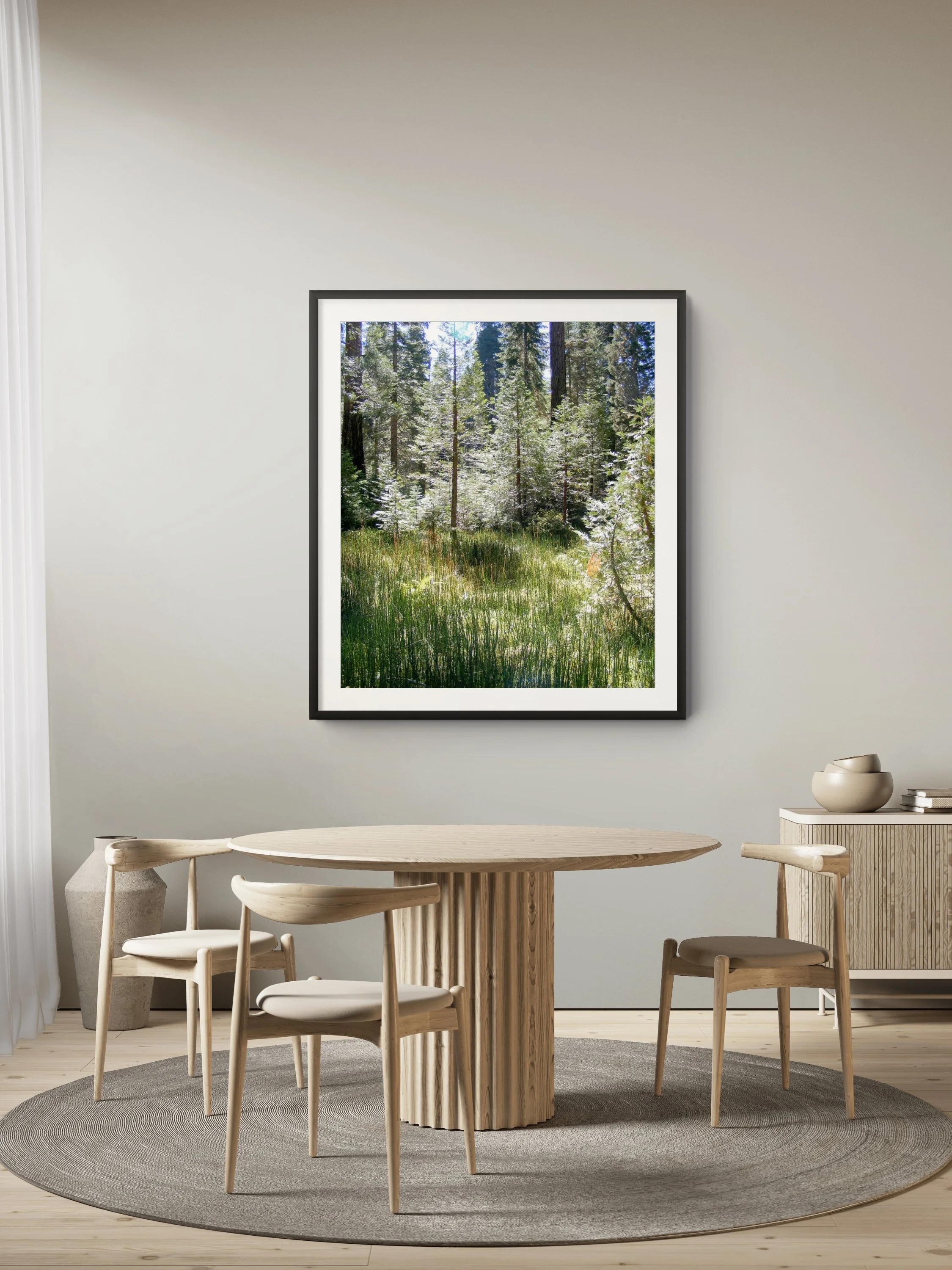 A dining room with a round wooden table, four wooden chairs with white seats, a grey circular rug, and a framed picture of a forest hanging on a light-colored wall.